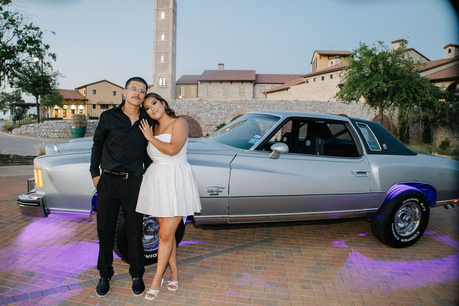 Couple in formal attire posing beside a classic silver lowrider car with purple underglow lighting at dusk.