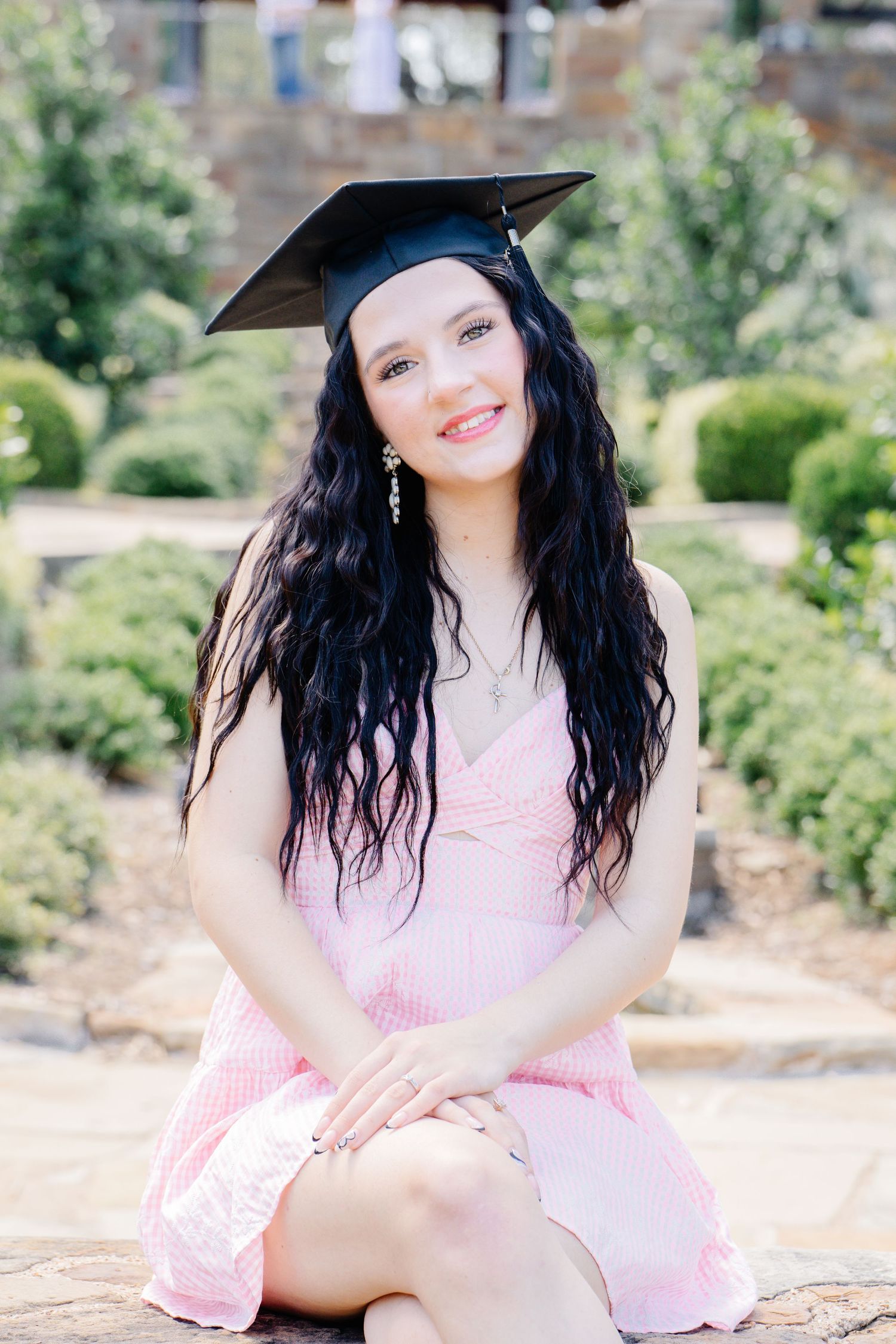 A smiling young woman wearing a graduation cap and pink dress sits outdoors in a lush green garden.