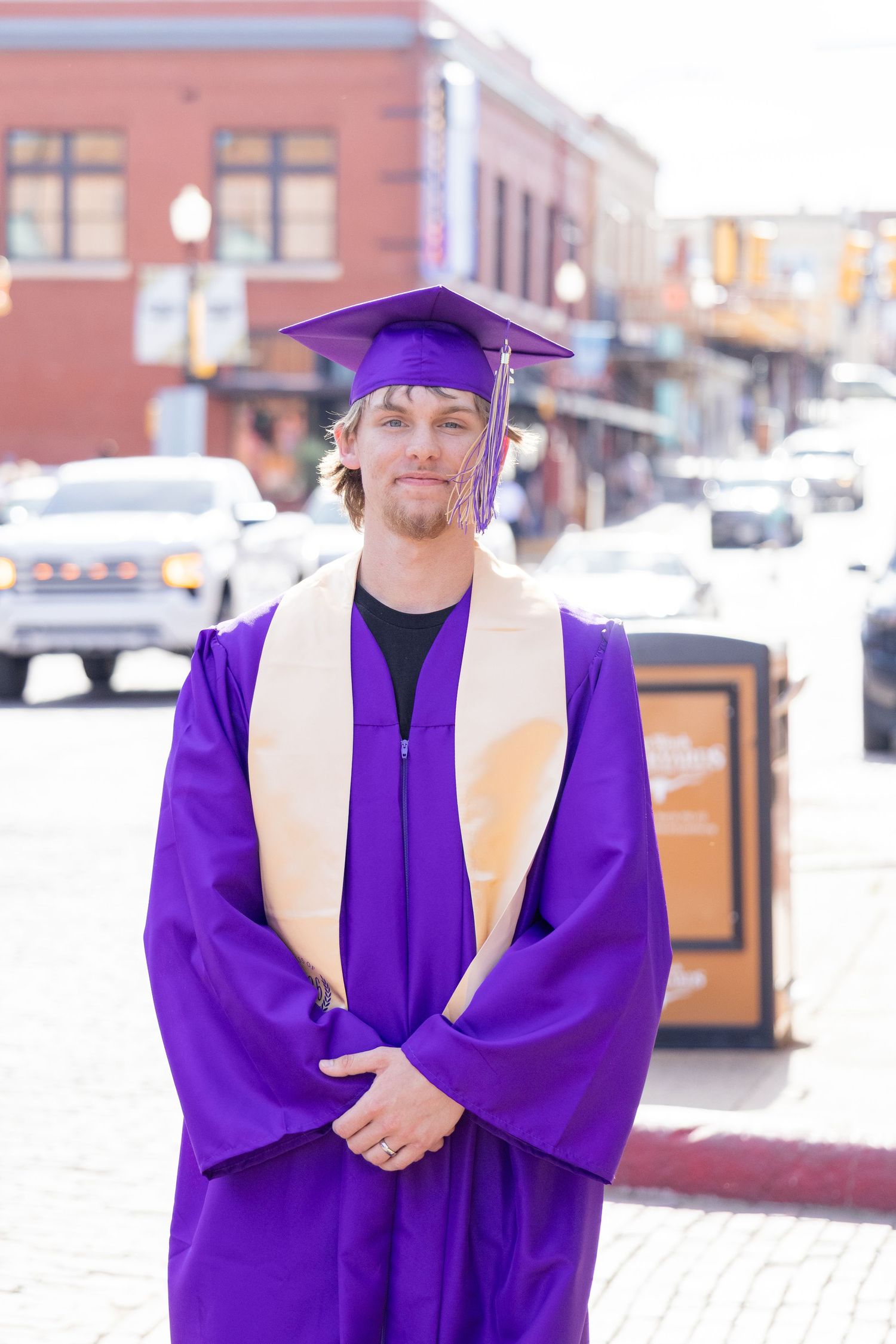 Young man in purple graduation cap and gown stands on brick street in urban setting.