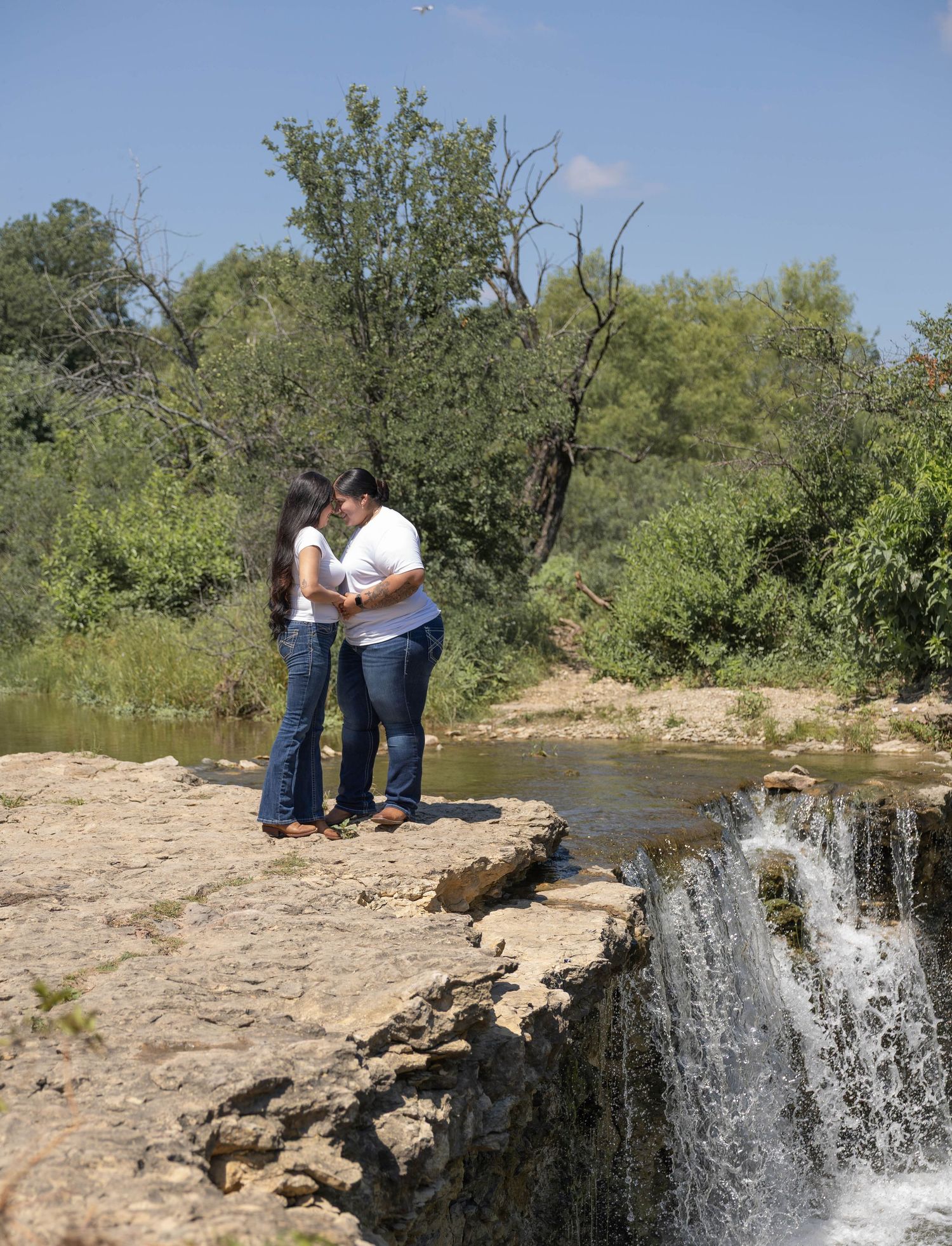 A couple stands close together on rocky ledge beside a cascading waterfall in a green landscape.