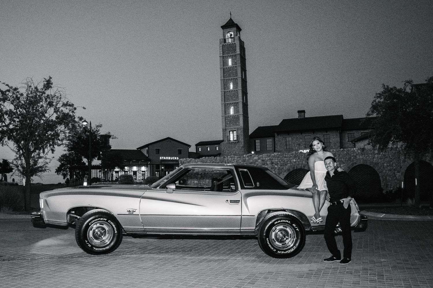 Black and white photo of a couple posing beside a classic muscle car with a clock tower in the background at dusk.