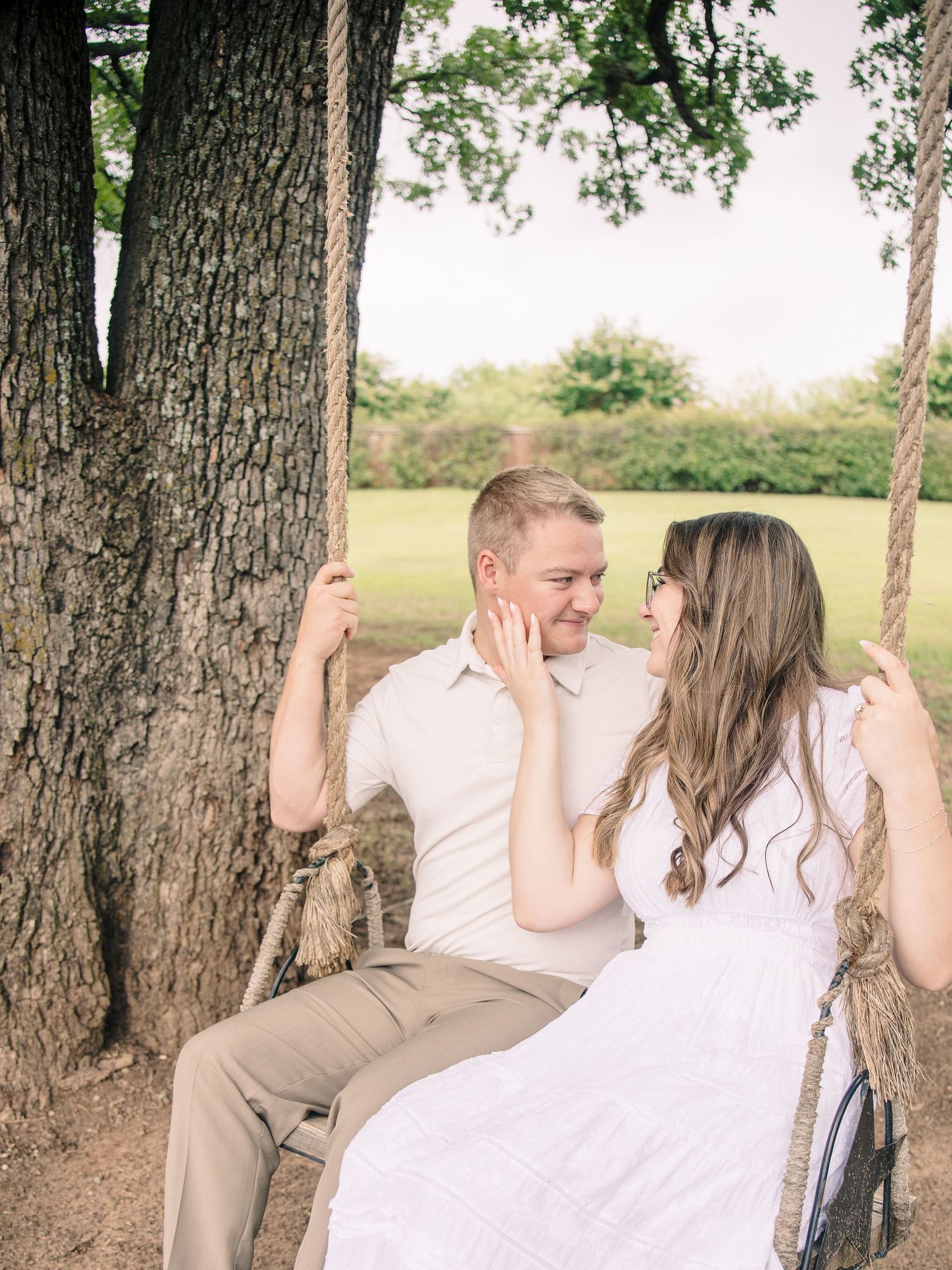 Couple sitting on a rope swing under a large tree, smiling at each other during an outdoor engagement photo session.