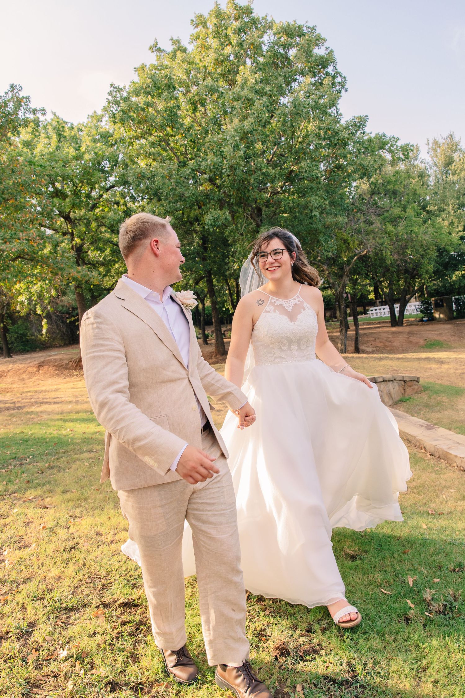 Smiling bride in a white lace gown and veil walks hand in hand with a groom in a tan suit across a sunny grassy lawn beneath tall trees.
