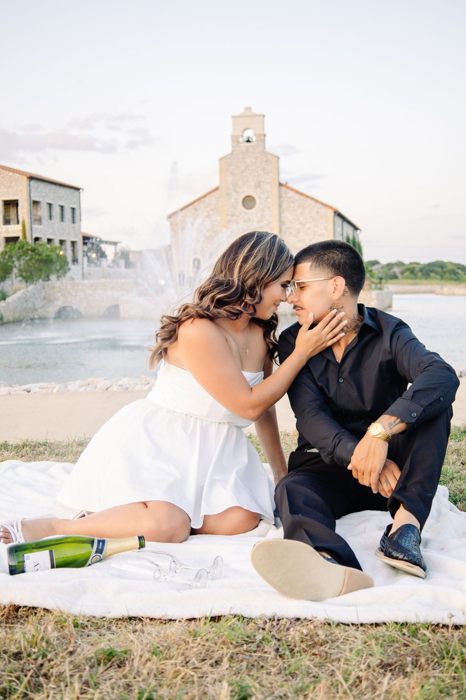 Engaged couple sharing a romantic moment on a blanket with champagne by a stone chapel.