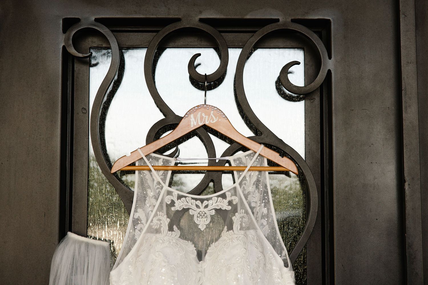 A lace wedding dress hangs on a wooden personalized hanger in front of an ornate wrought iron door.
