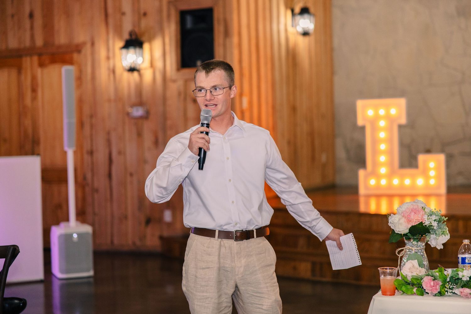 Man in glasses and a light button-up shirt gives a wedding toast into a microphone beside a marquee letter sign and floral centerpieces.
