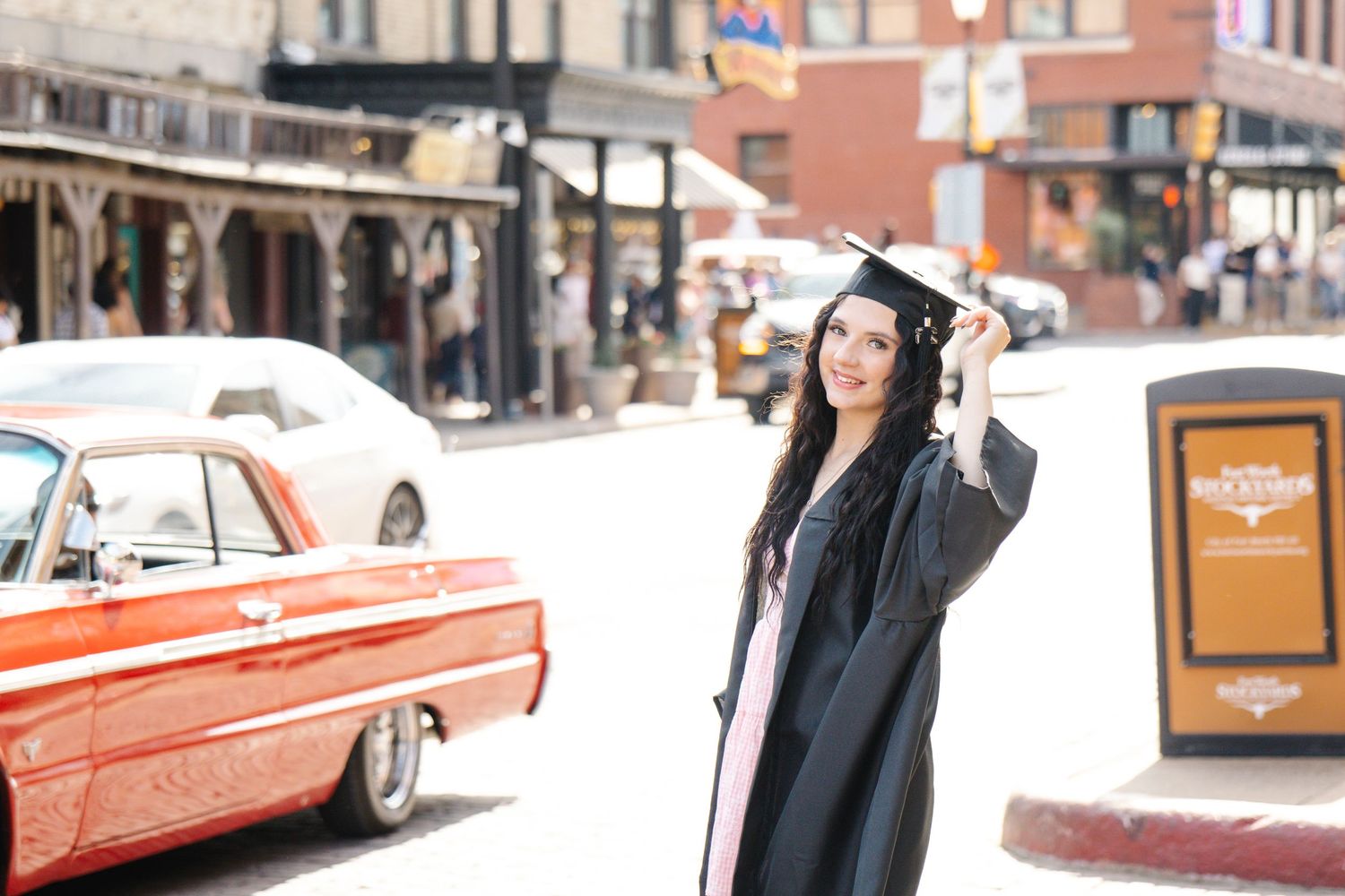 Smiling graduate in a black gown and cap poses on a sunny downtown street beside a vintage red car and brick buildings. FTW Stockyards