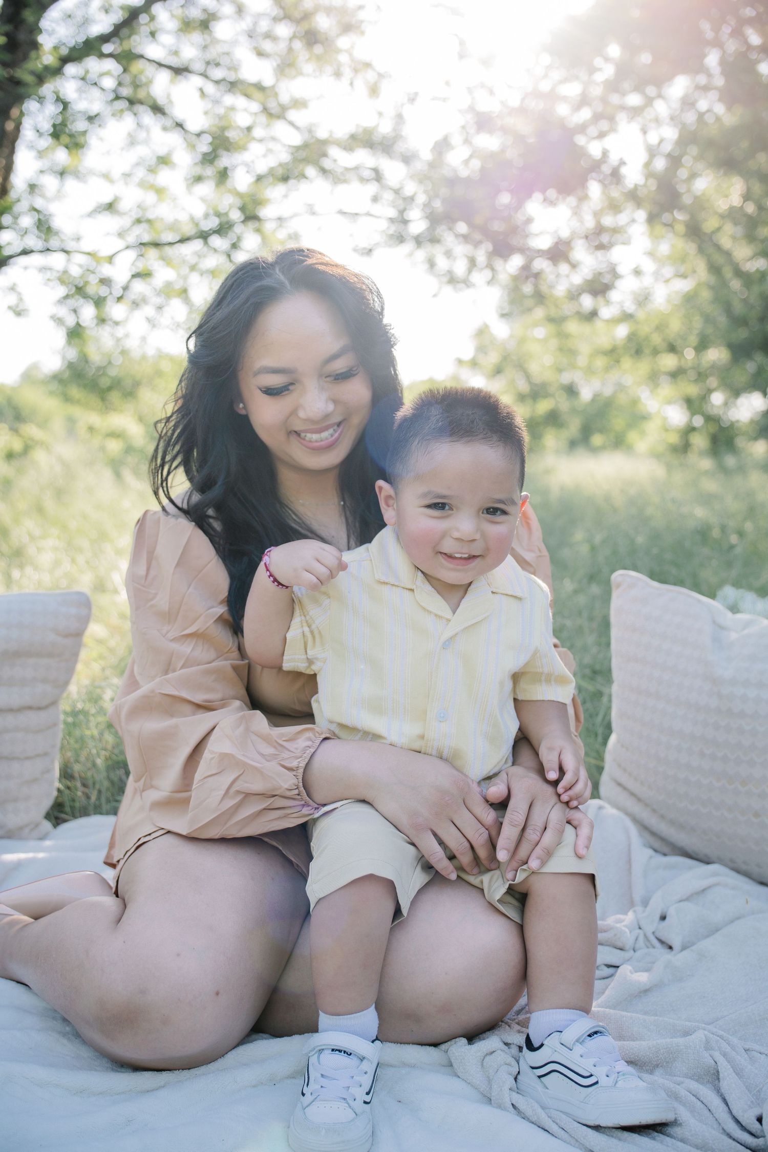 A smiling woman sits outdoors on a blanket with a young boy on her lap, surrounded by greenery in soft sunlight.