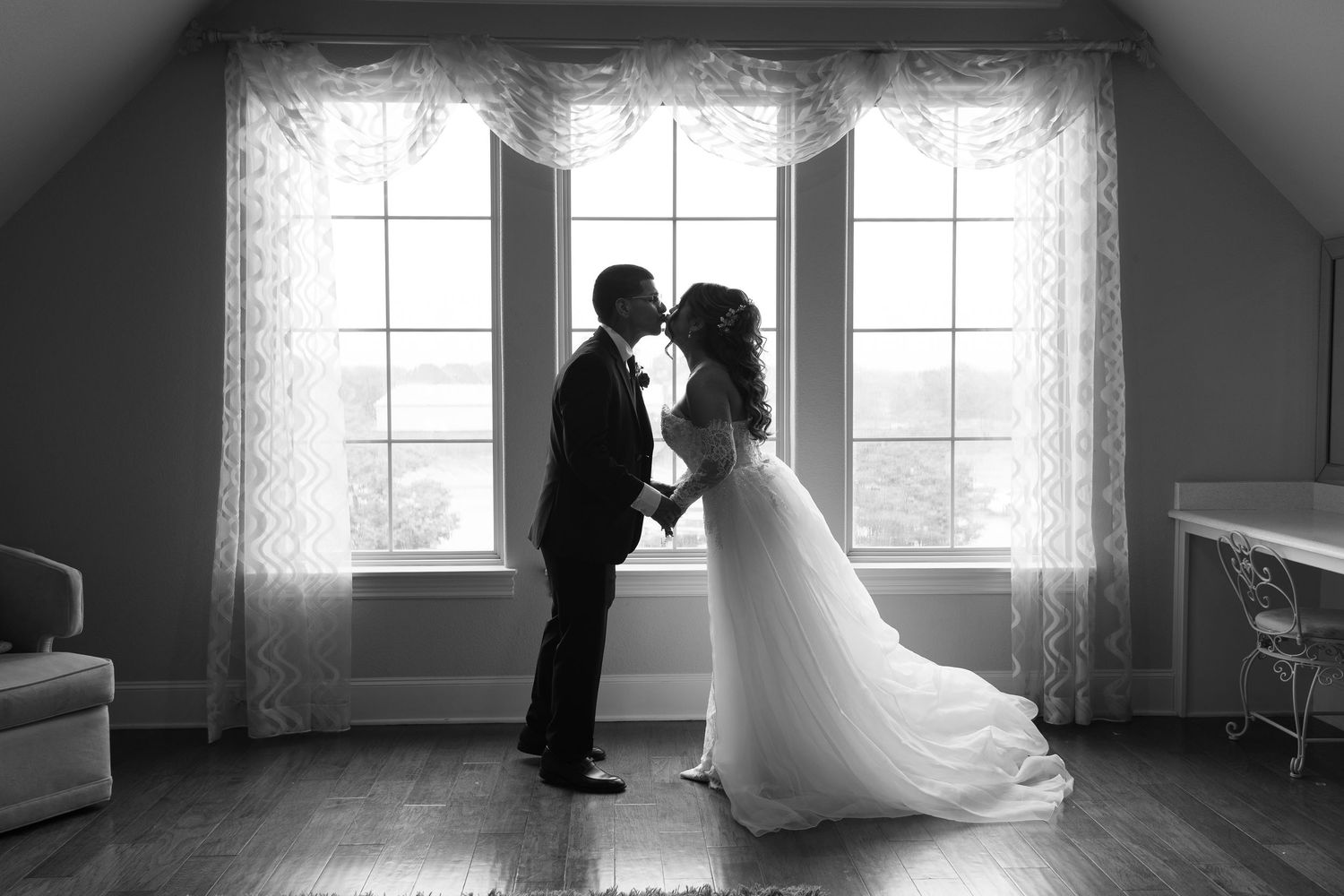 Black and white photo of a bride and groom sharing a kiss in front of large windows on their wedding day.