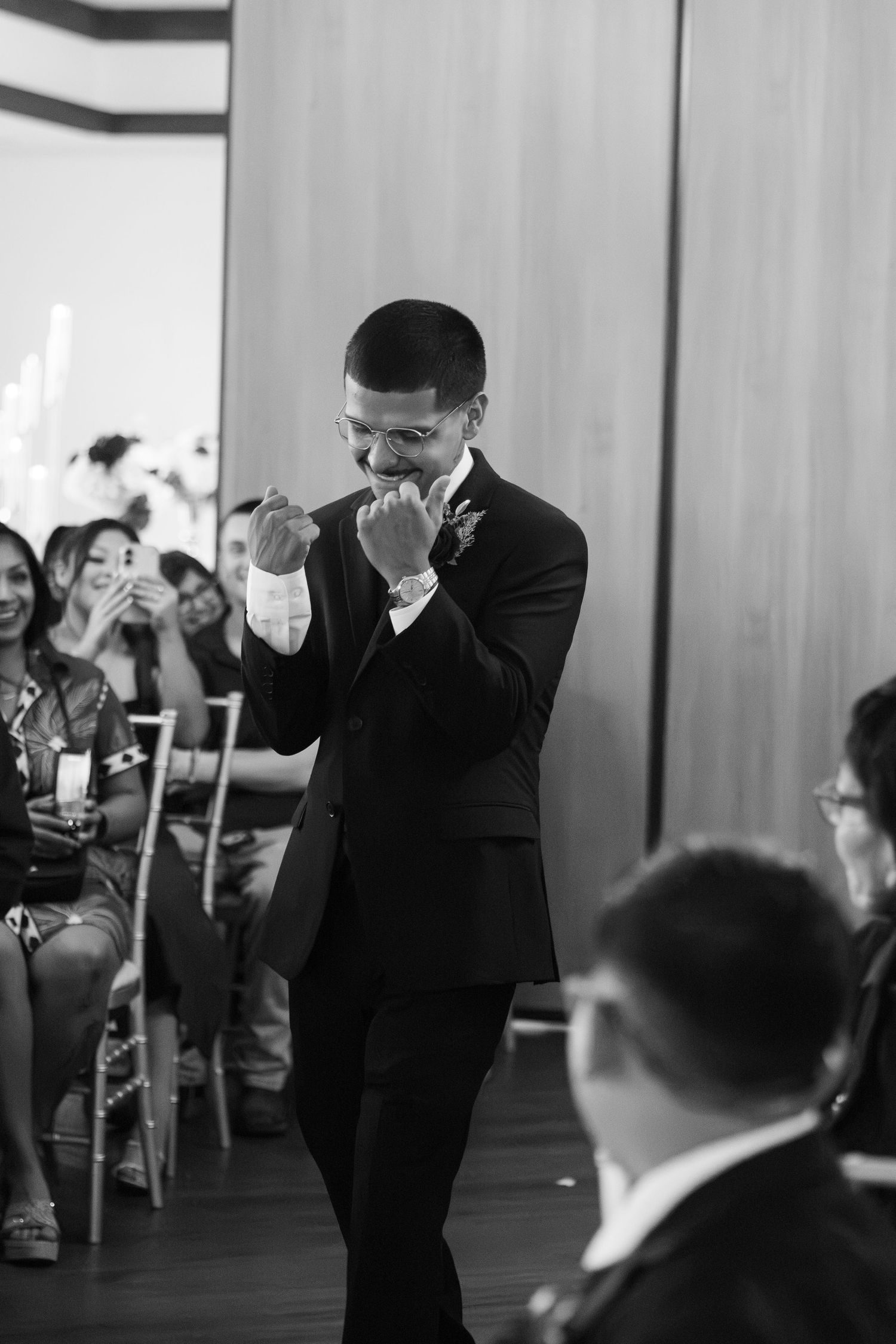 Emotional groom wipes tears in black and white photo as guests cheer during wedding ceremony.