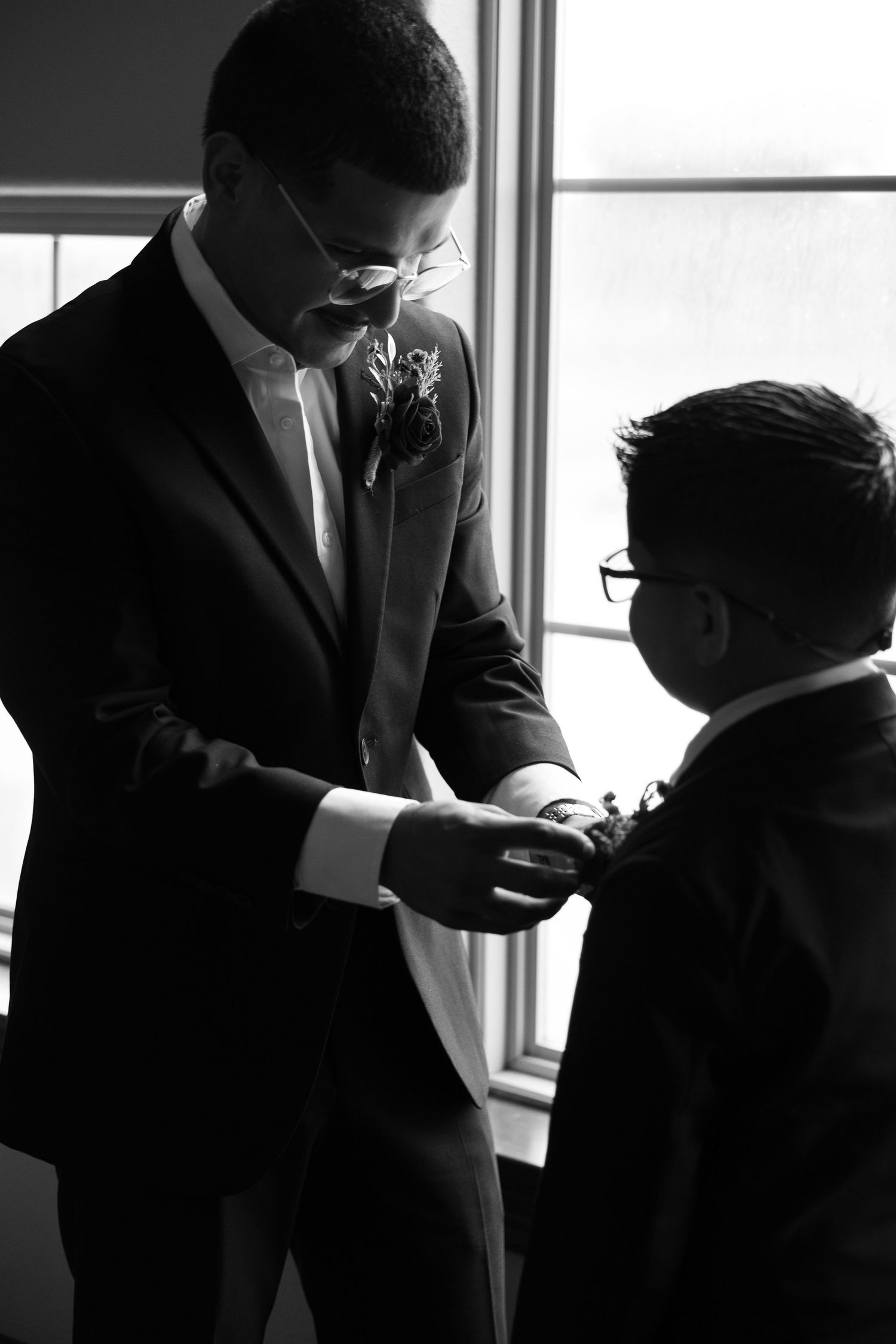 Groom in black tuxedo helps young ring bearer with his boutonniere in a black and white wedding photo.