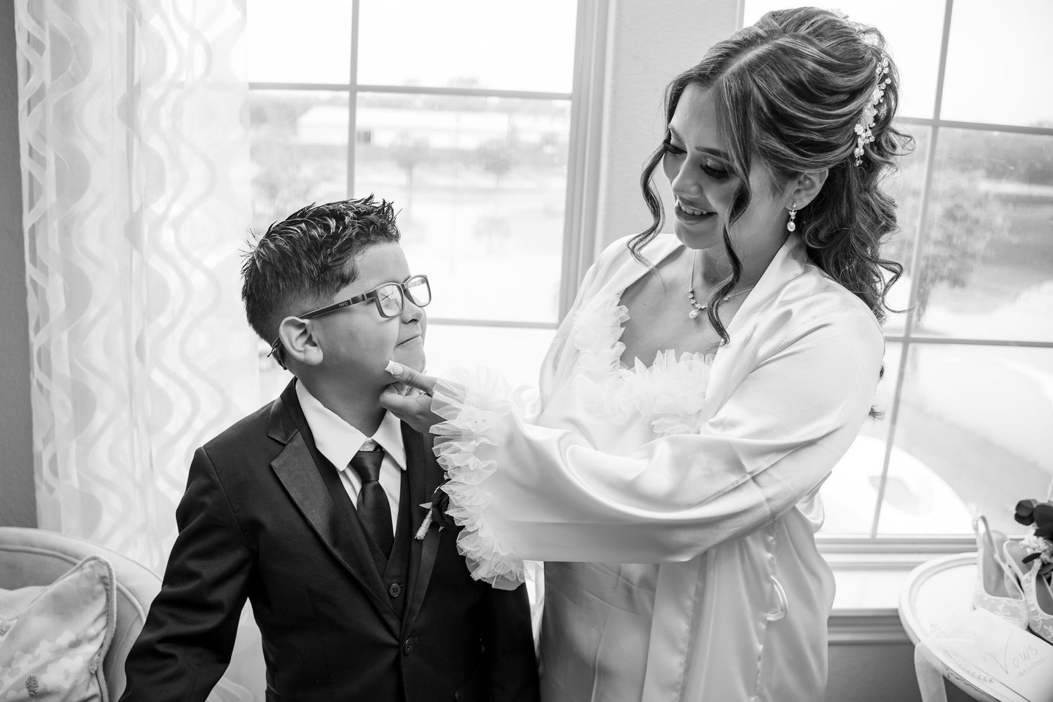 Bride in white robe adjusts young boy's tie near window in black and white wedding preparation photo.