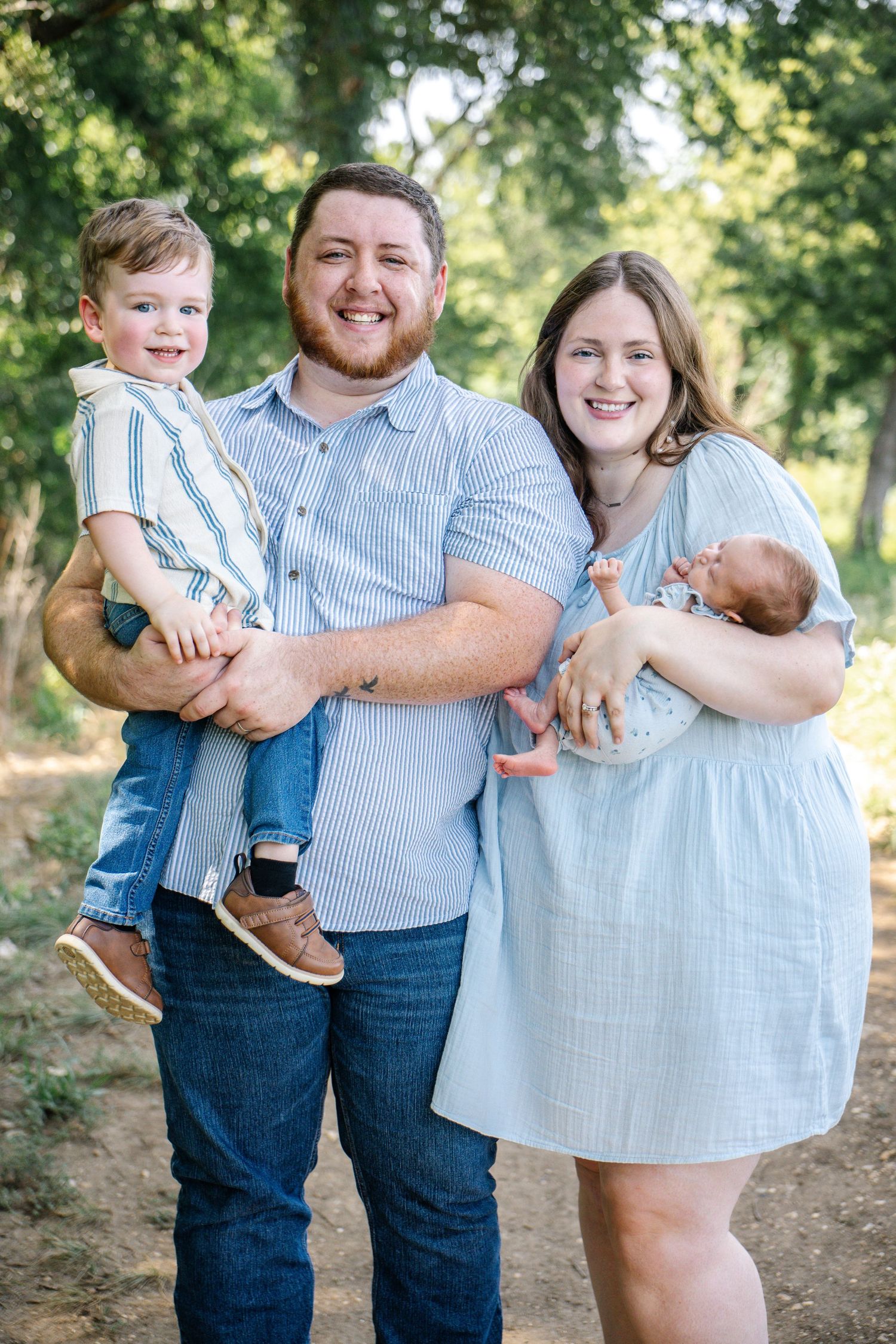 Happy family of four posing outdoors, parents holding two young children, dressed in coordinating blue outfits among green trees.