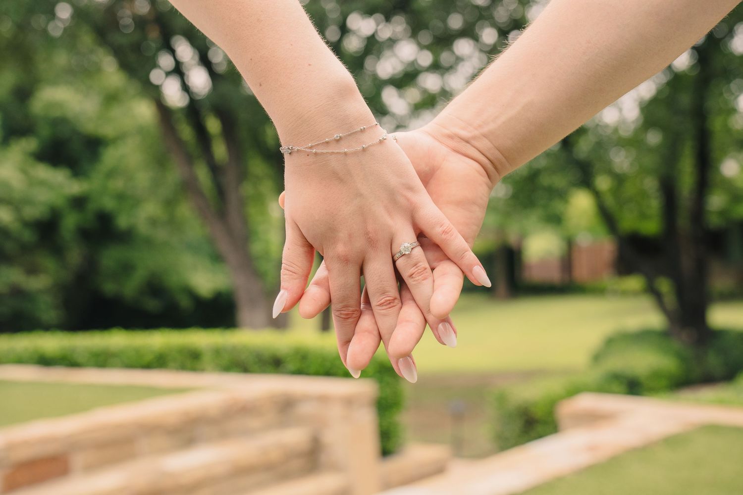 Two people holding hands outdoors, one wearing an engagement ring and bracelet, with a lush green garden background.