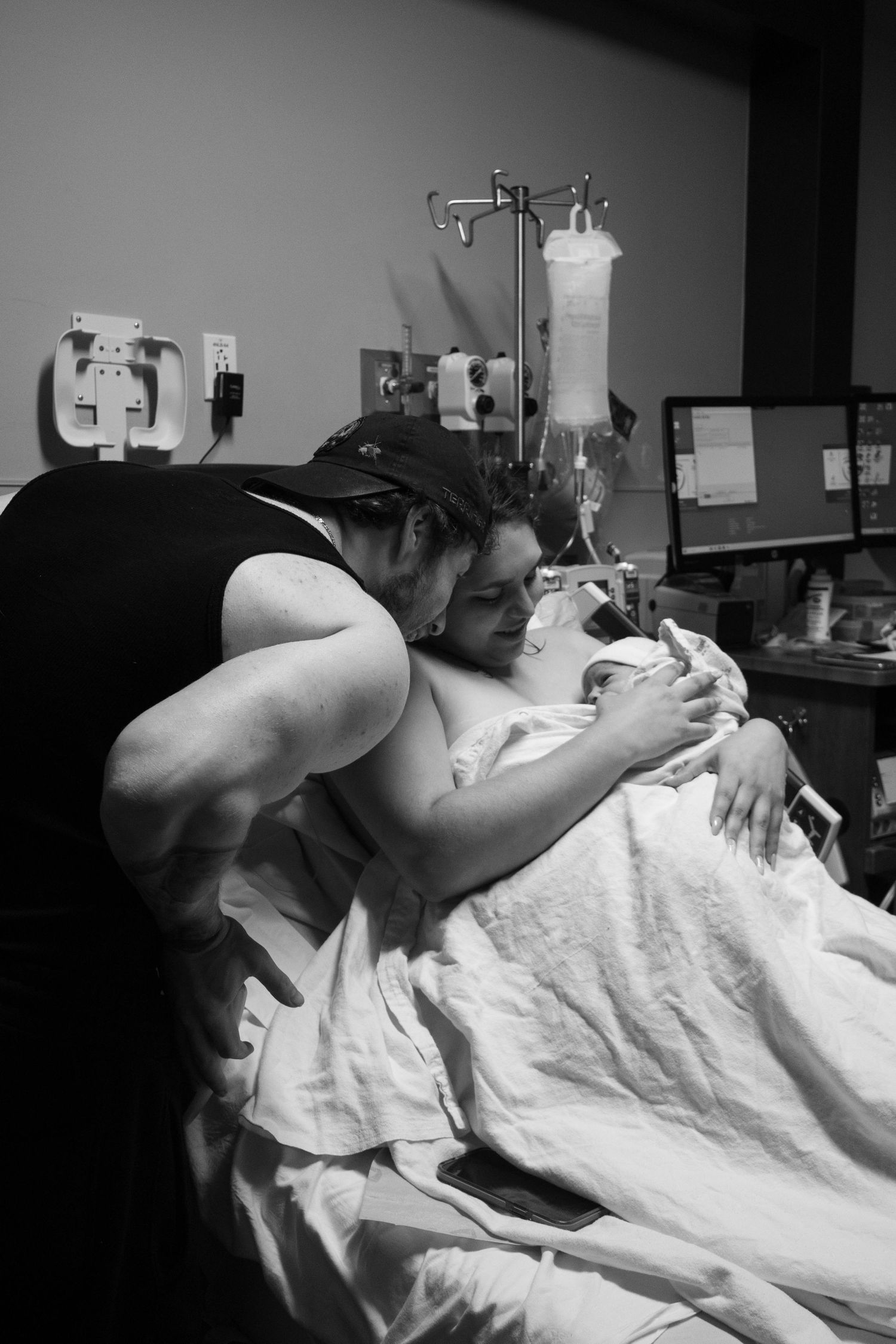 A couple shares a tender moment in a hospital room after childbirth, with the mother resting in bed holding their newborn.