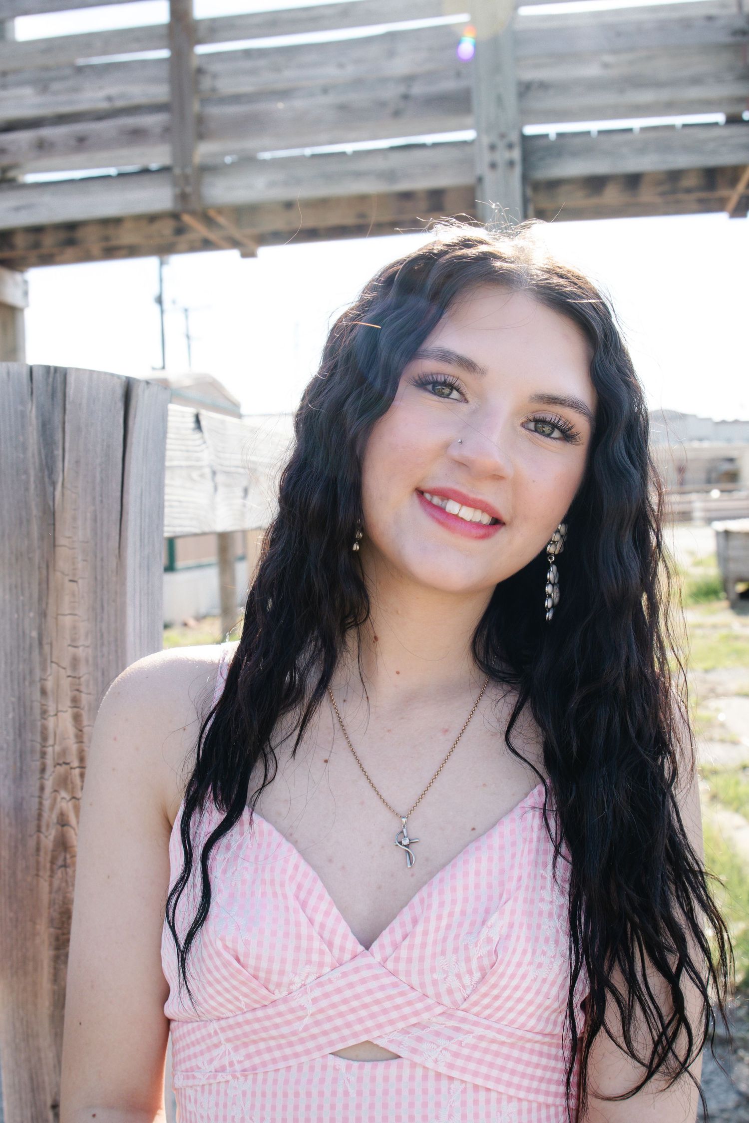 Young woman with long dark hair smiling outdoors in a pink top with crystal earrings and necklace.
