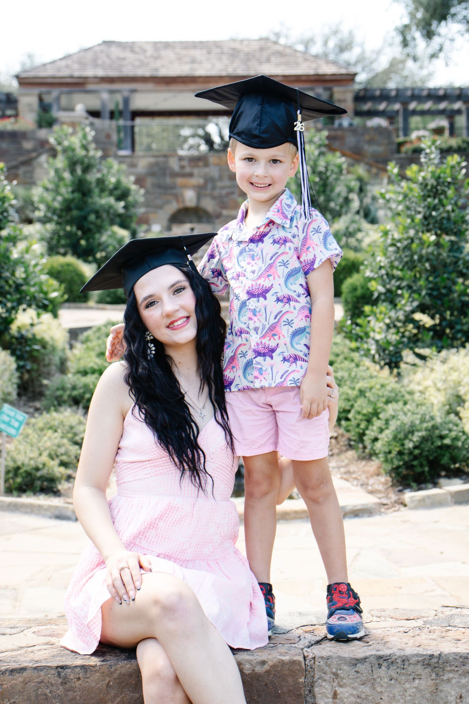 Woman in pink dress and graduation cap poses with young boy in colorful shirt outdoors.