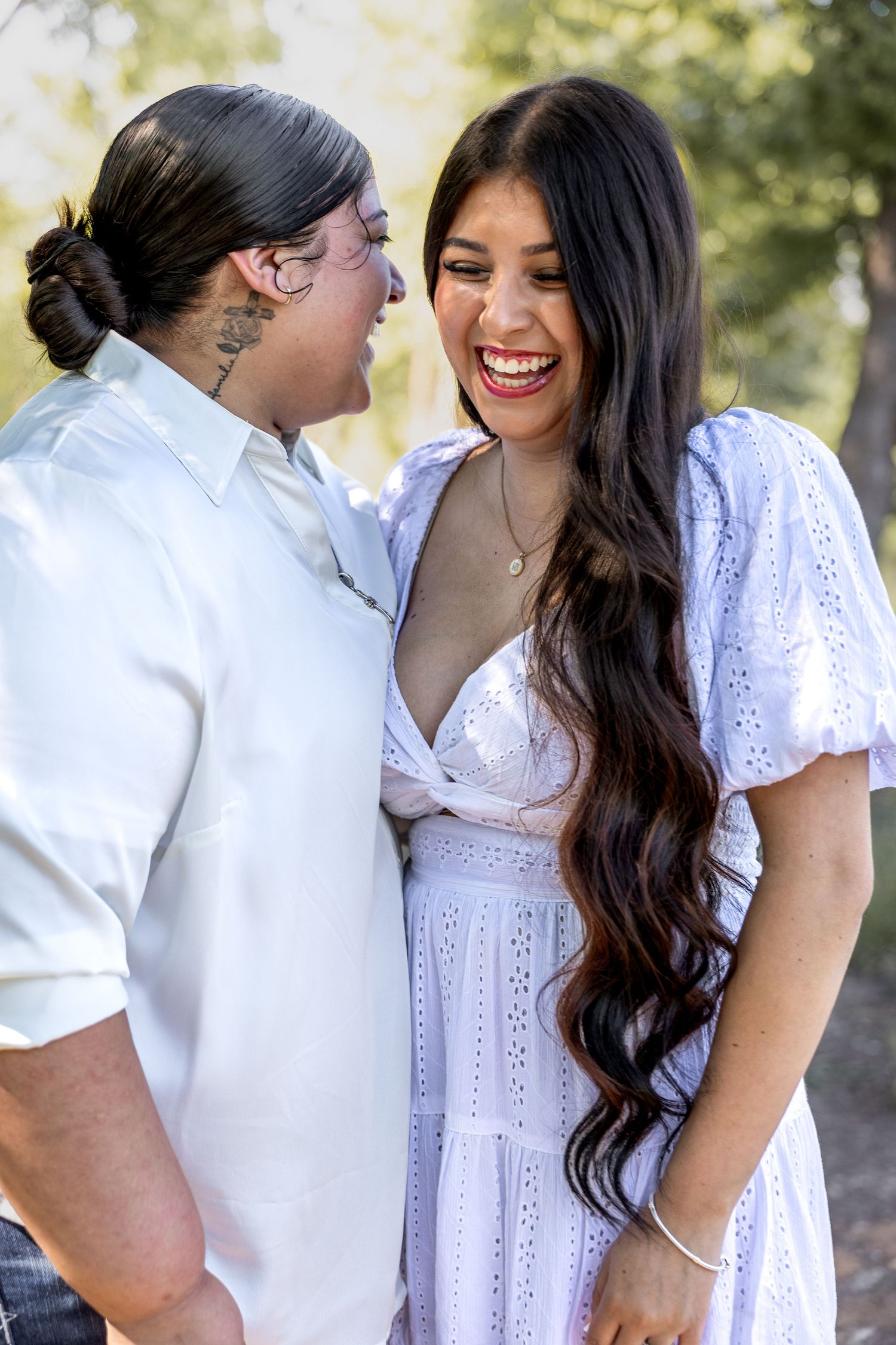 Two women laughing together outdoors in white outfits in a sunlit park.