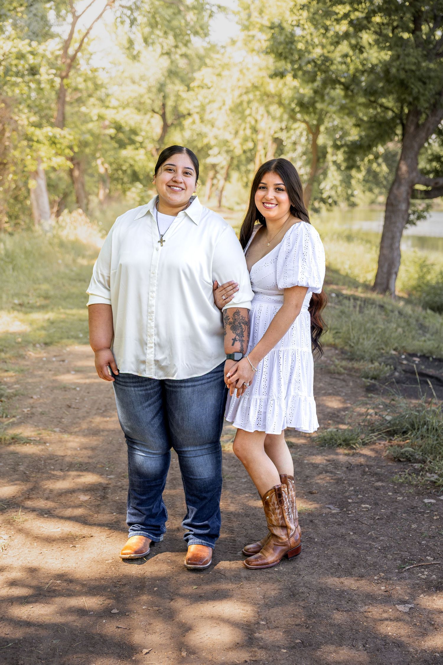 Smiling couple standing together on a shaded dirt path, woman in white dress with cowboy boots holding hands.