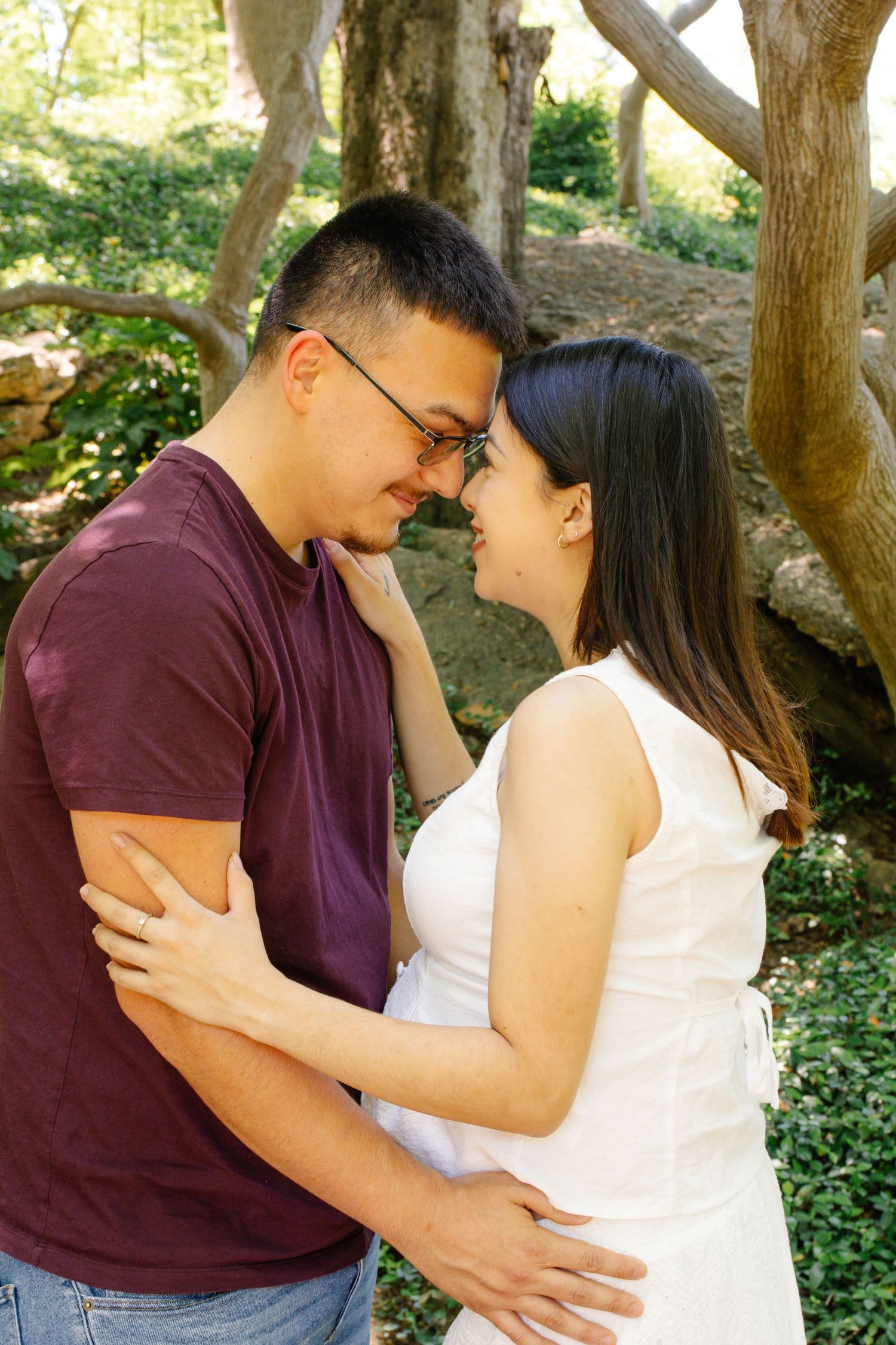 Couple sharing a tender forehead-to-forehead moment surrounded by lush green trees in a park.