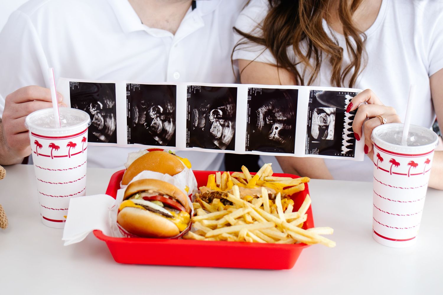Couple holding ultrasound photos over In-N-Out Burger tray with burgers and fries on the table.