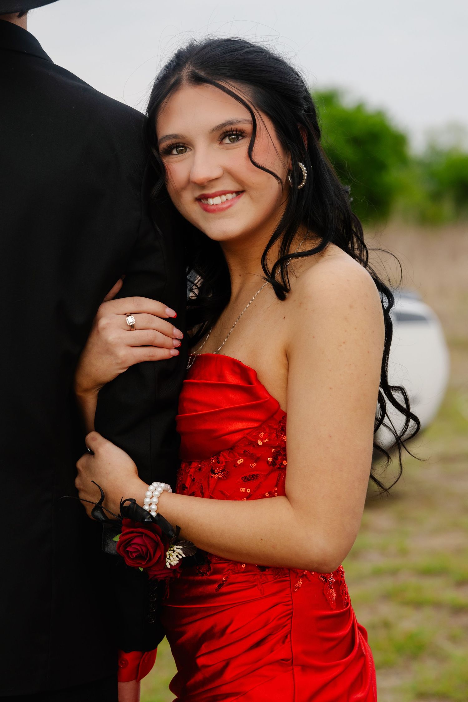 Smiling young woman in red strapless gown with corsage embracing her prom date outdoors.