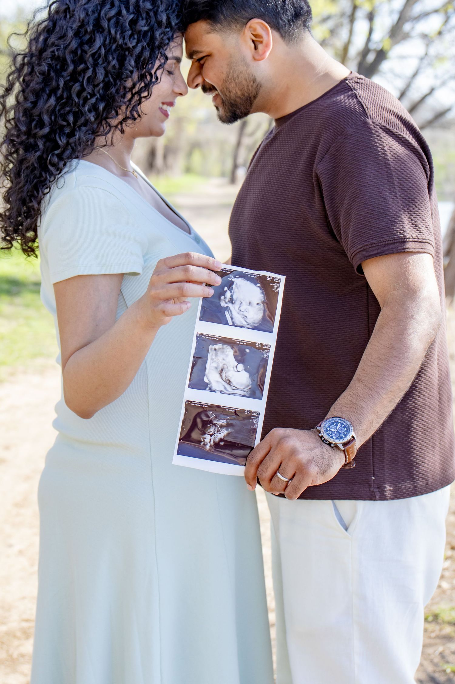 Expecting couple holding ultrasound photos while touching foreheads lovingly in a sunny outdoor park setting.