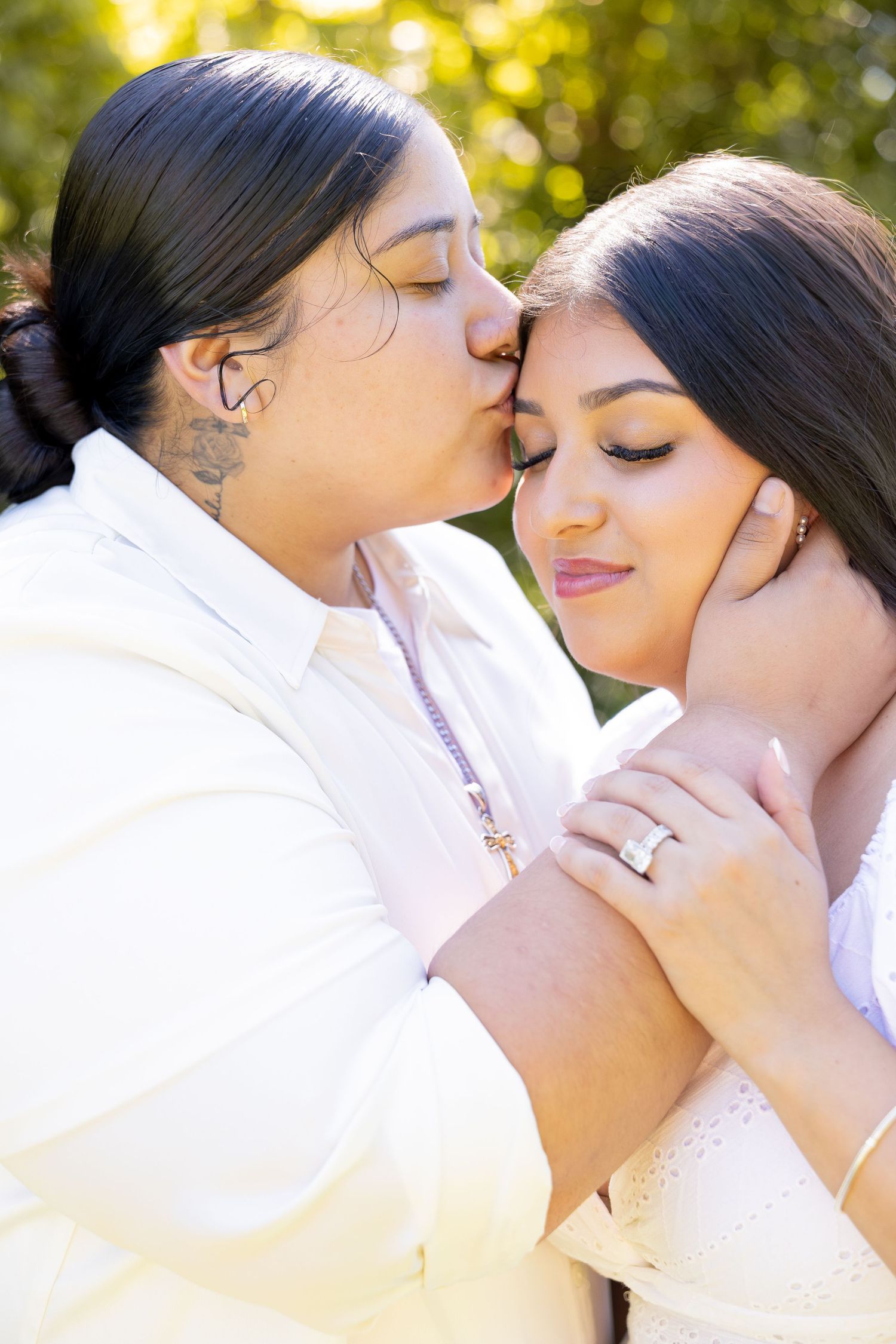 Two women share a loving kiss on the cheek while embracing closely in a sun-drenched outdoor setting.