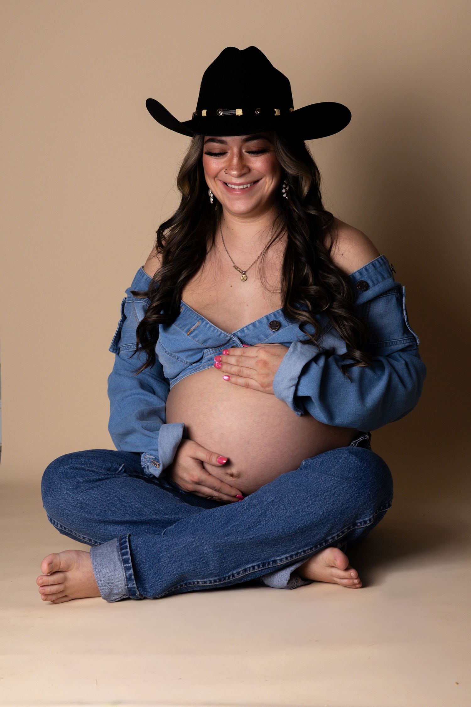 Smiling pregnant woman in denim outfit and black cowboy hat sitting cross-legged on studio floor.