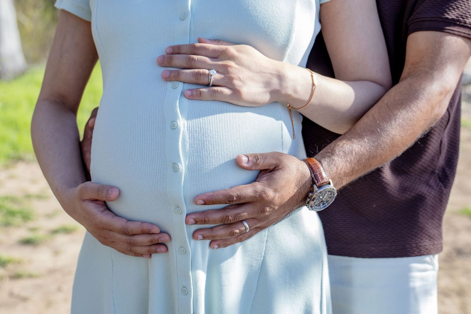 Couple gently cradling pregnant belly outdoors, man wearing watch embracing woman in white dress.