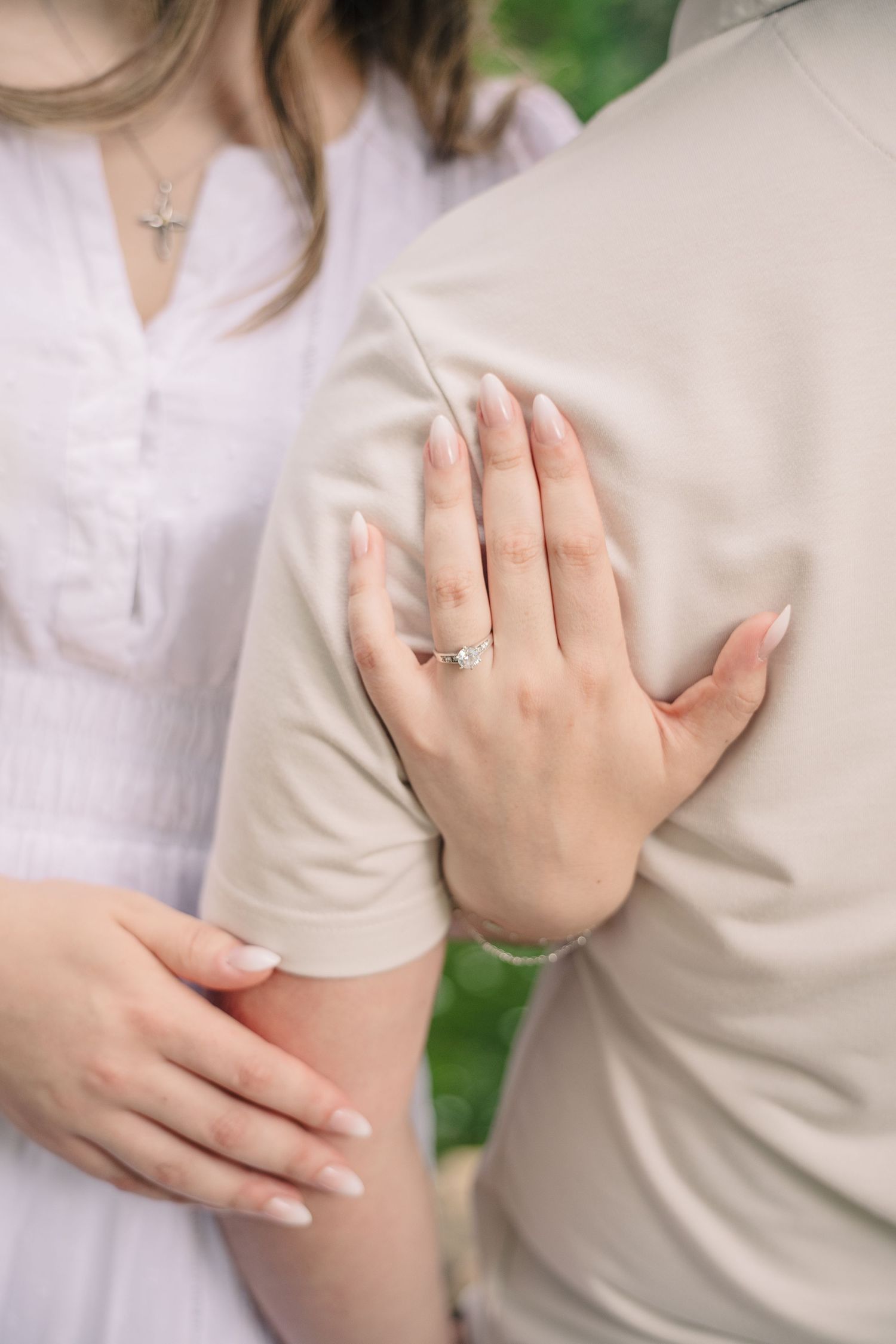 Close-up of womans hand showing diamond engagement ring while embracing her partner outdoors.