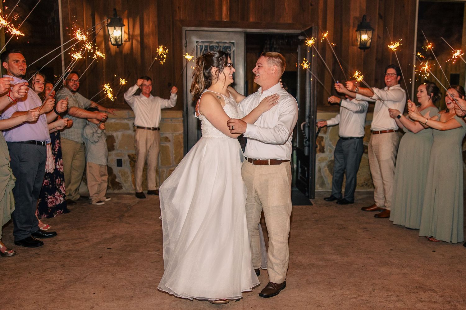 Bride and groom dance close together as wedding guests hold lit sparklers during a festive evening send-off.