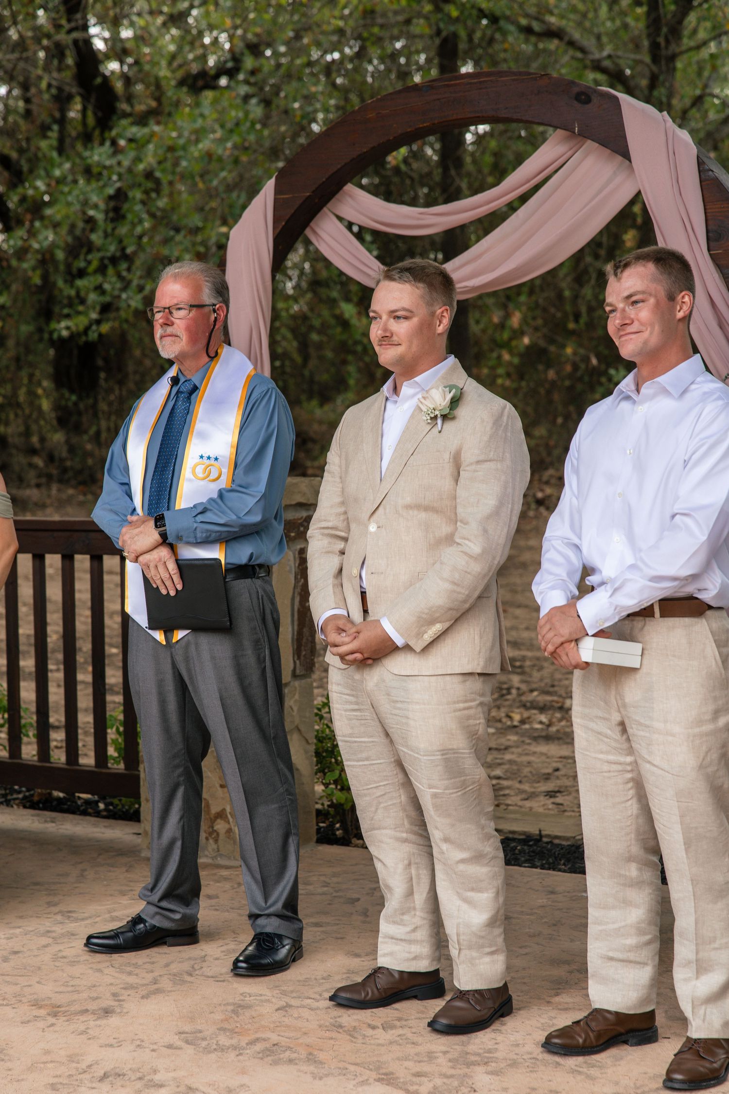 Groom in a tan linen suit stands smiling between an officiant in a stole and a groomsman beneath a draped wedding arch.
