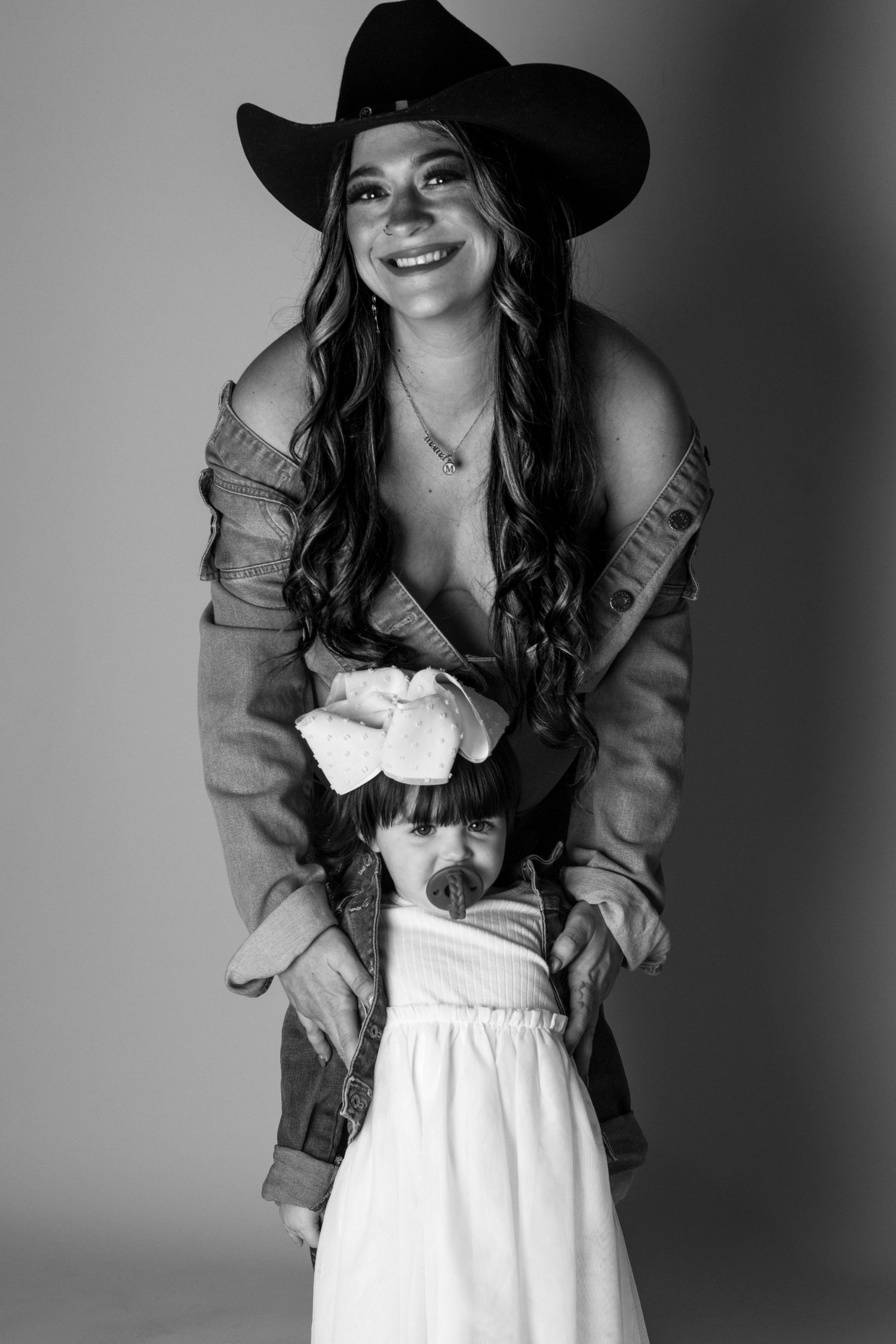 Black and white photo of a smiling woman in cowboy hat and denim jacket posing with a young girl in white dress.