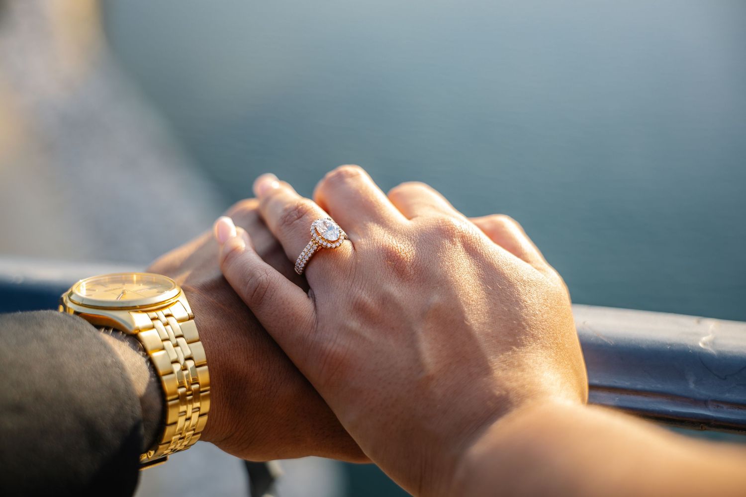 Close-up of couple holding hands, woman wearing diamond engagement ring, man wearing gold watch, outdoor setting.