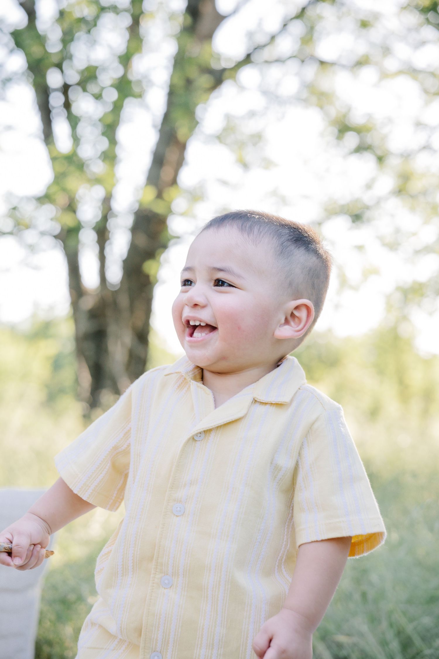 Happy toddler boy in yellow striped shirt smiling outdoors with green trees in background.