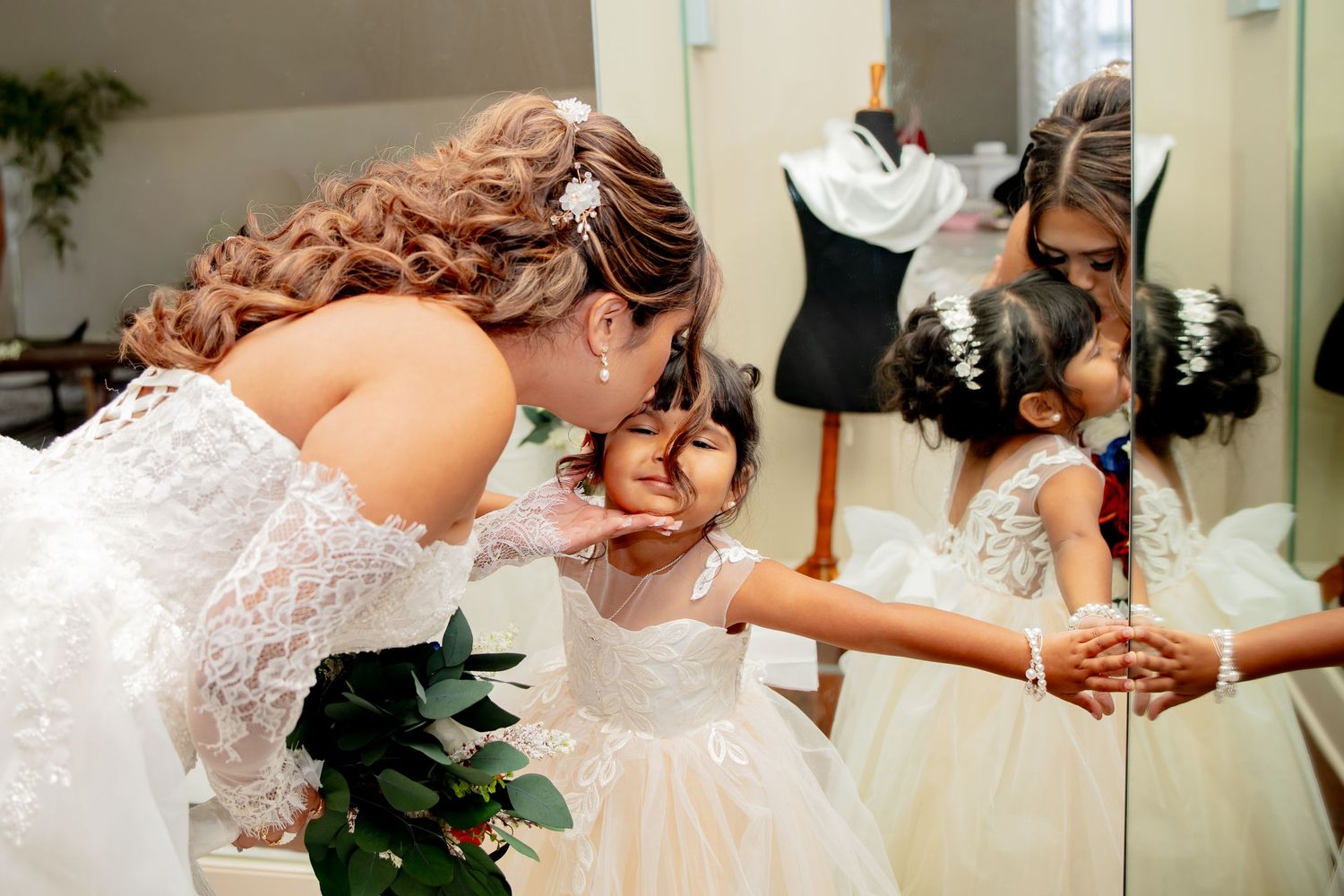 Bride in white lace gown kissing young flower girl in tulle dress, reflected in mirror at wedding venue.