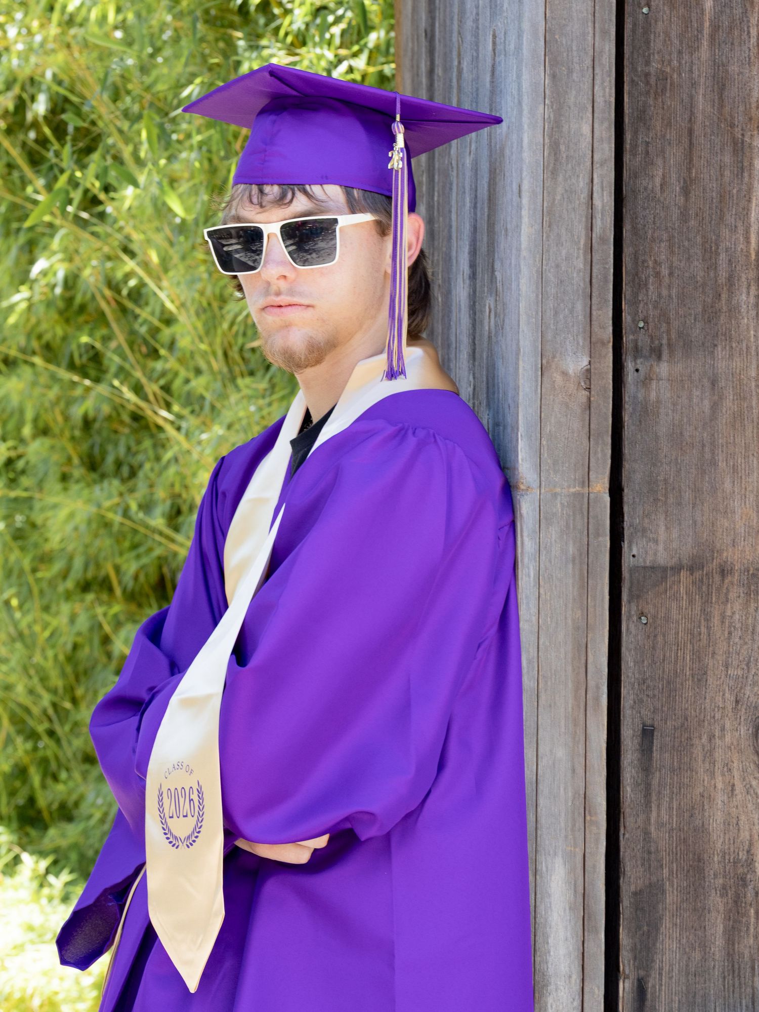 Graduate in purple cap and gown wearing sunglasses leans against rustic wooden wall outdoors.