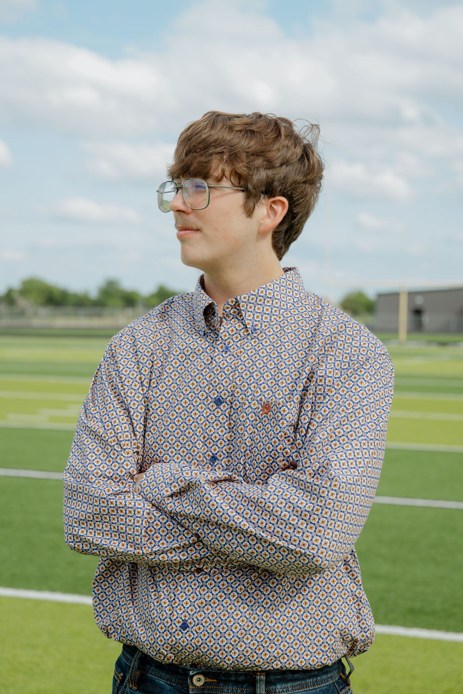 Young man in patterned button-down shirt with arms crossed gazing away on a green sports field.