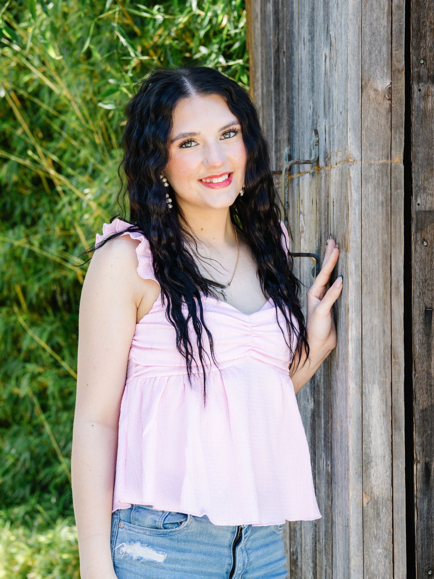 A young woman with dark curly hair smiles in a pink top leaning against a rustic wooden door.