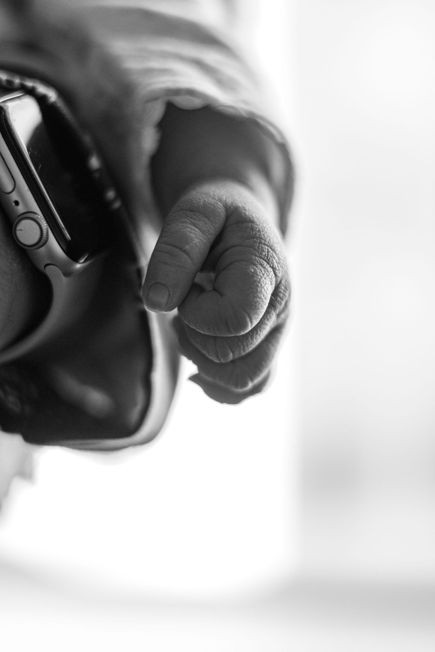 Black and white macro photo of a newborn baby's tiny fist next to parent's smartwatch.