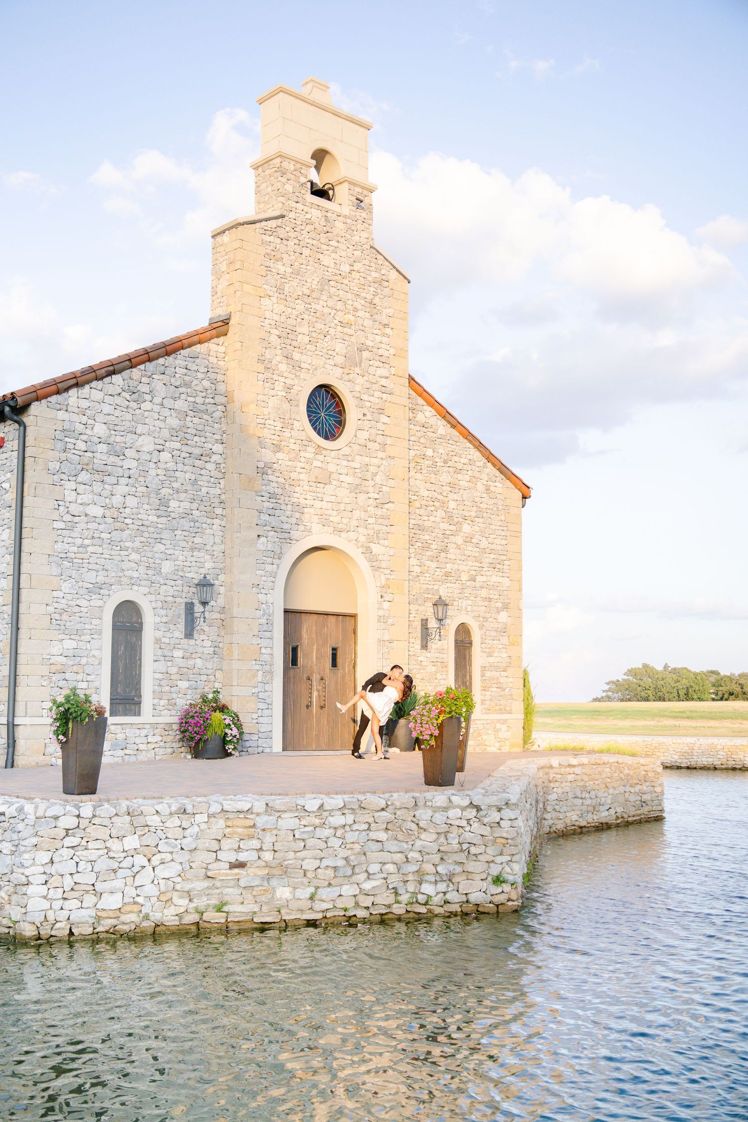 Couple kissing on steps of rustic stone chapel by water at golden hour during outdoor wedding ceremony.