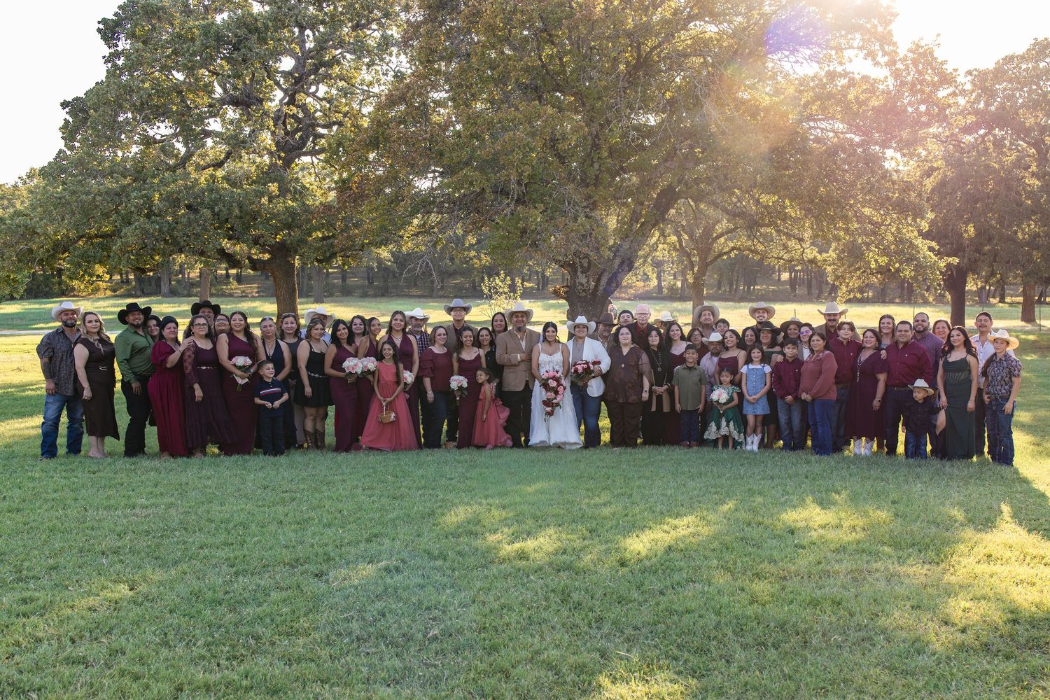 Large group of people in formal burgundy and dark attire posing together on a sunny grass lawn beneath tall shady trees at an outdoor event.