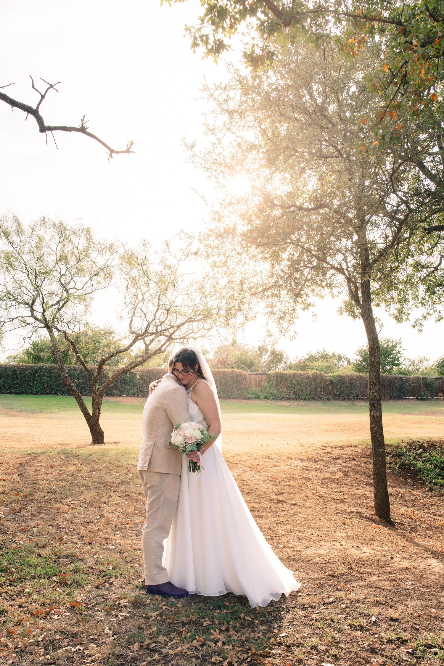 Bride in a flowing white gown holds a bouquet and embraces her groom in a tan suit amid golden-hour trees.
