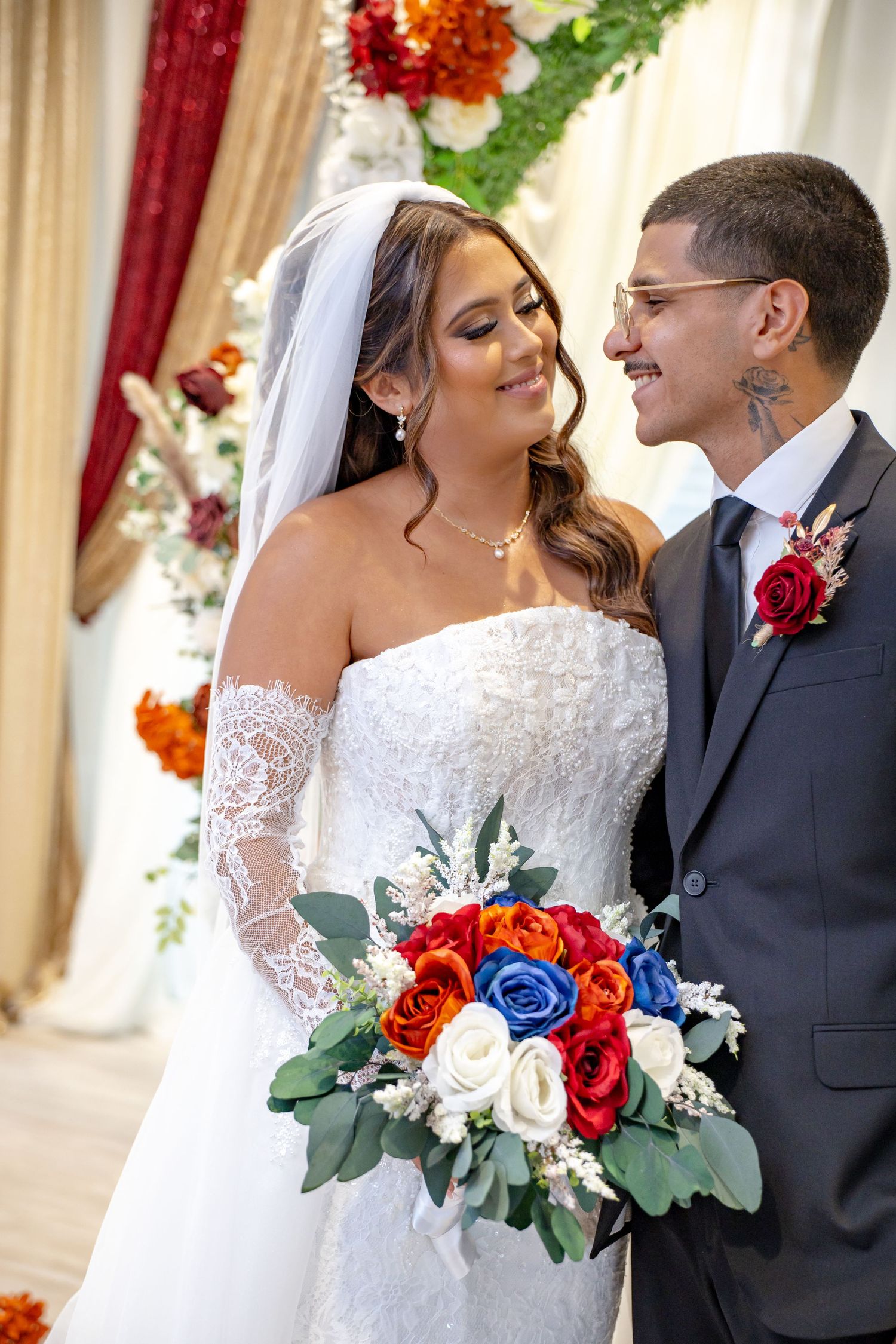 Bride and groom share a loving glance during ceremony, bride holding vibrant red, blue, and white bouquet.