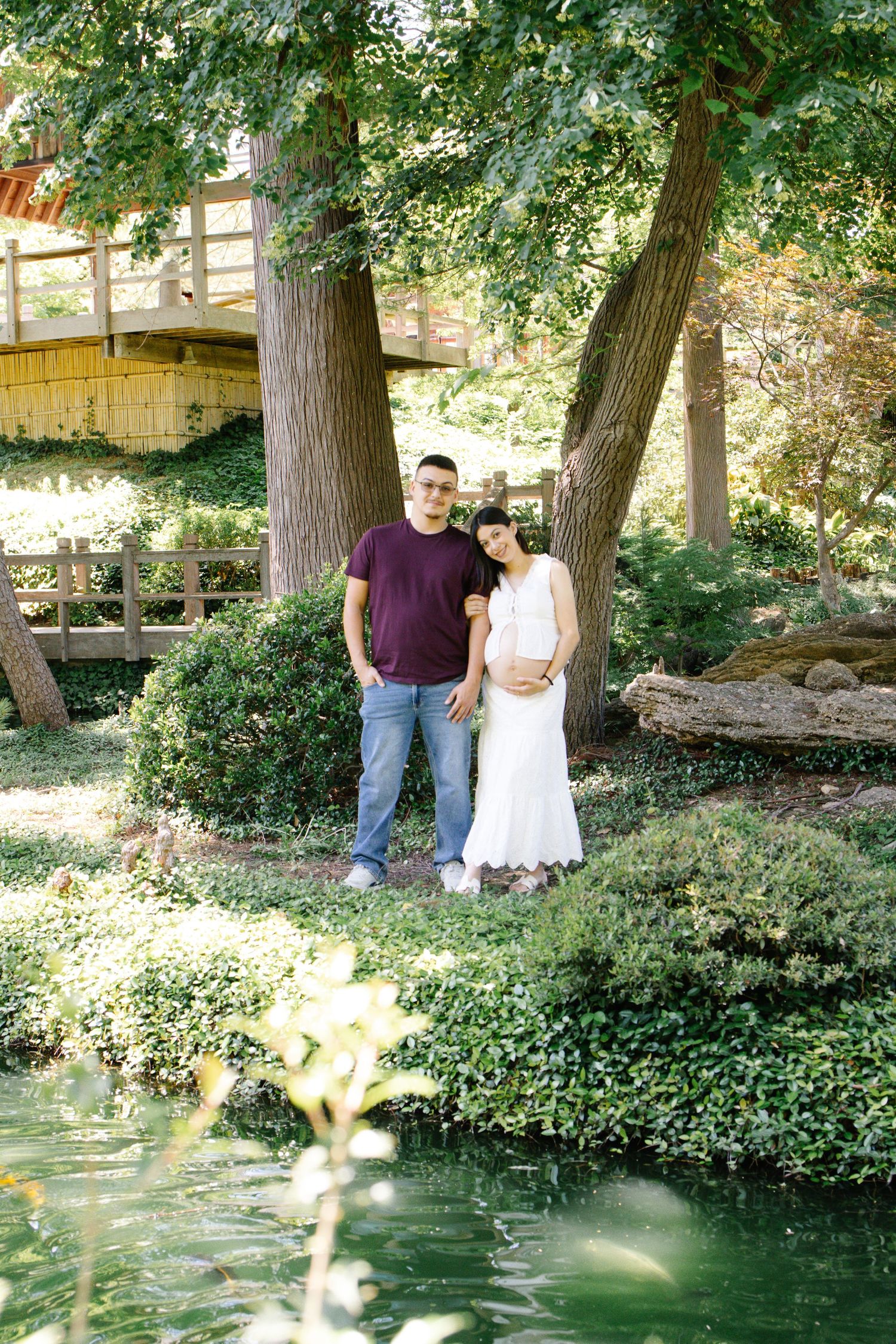 Expecting couple standing together near a serene pond surrounded by tall trees and greenery.