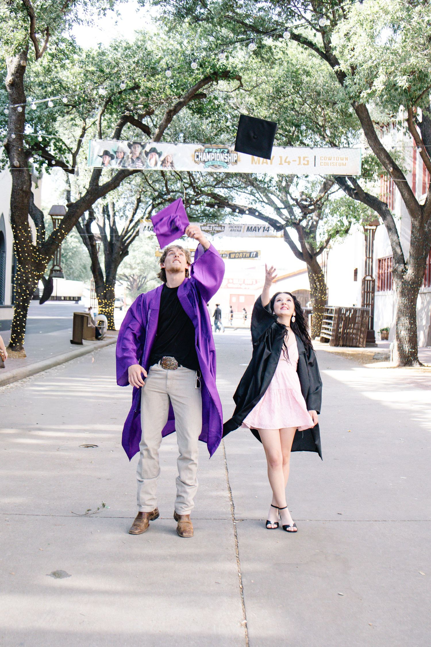 Two graduates in purple and black gowns tossing caps on a tree-lined street.
