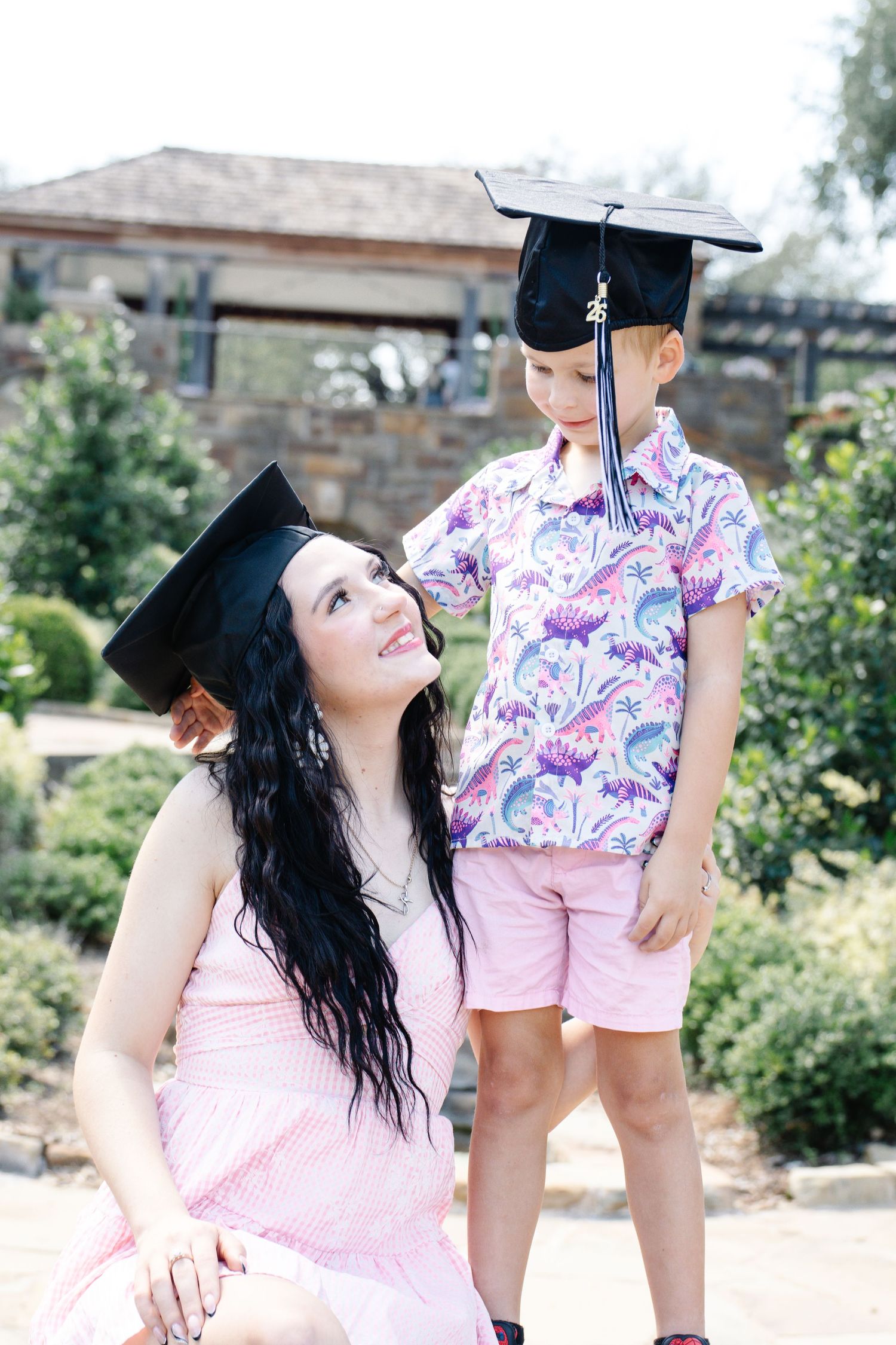 Woman in graduation cap and pink dress smiling at young boy wearing graduation cap outdoors.