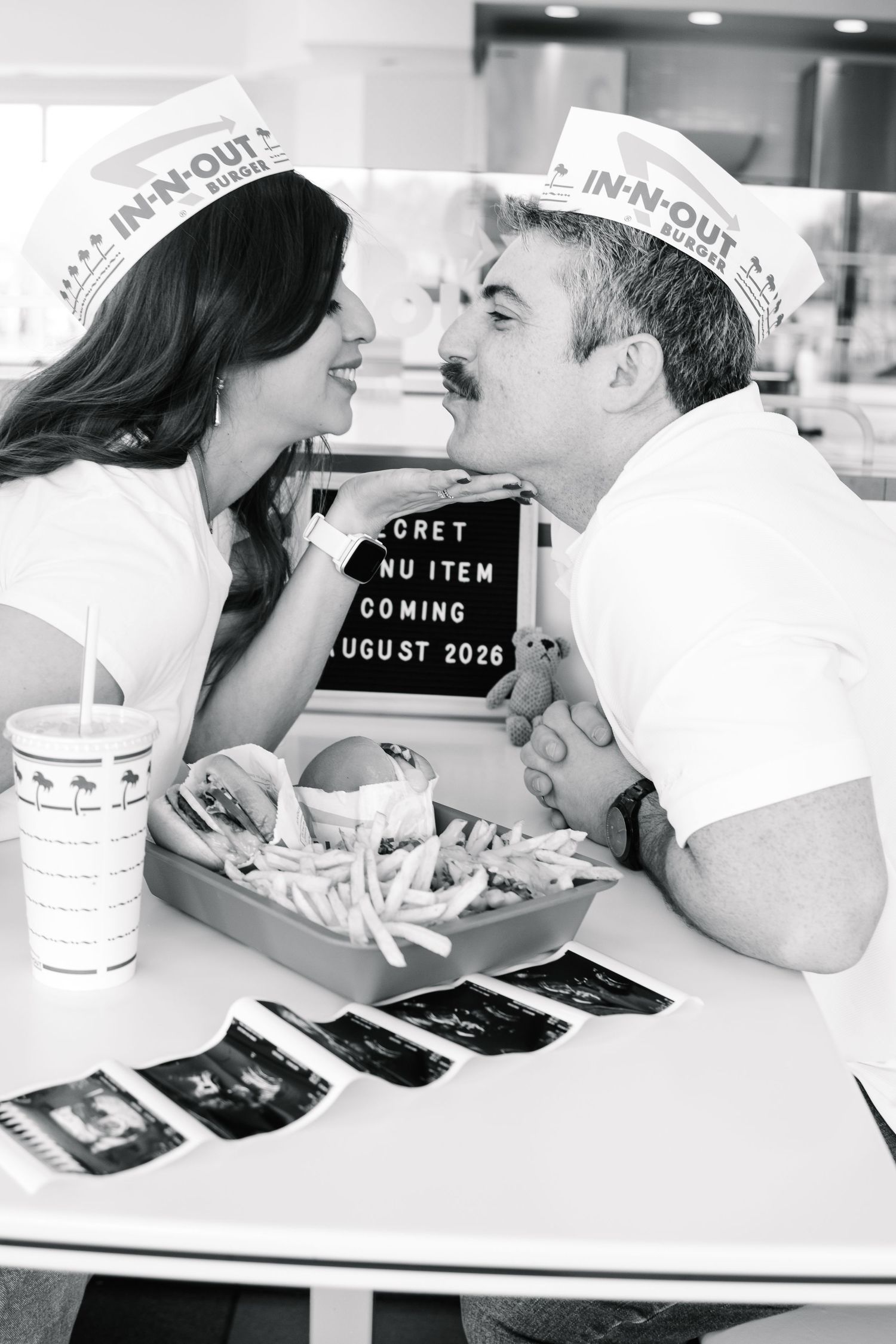 Couple kissing at In-N-Out Burger with a letter board announcing a secret menu item coming August 2026 and ultrasound photos.