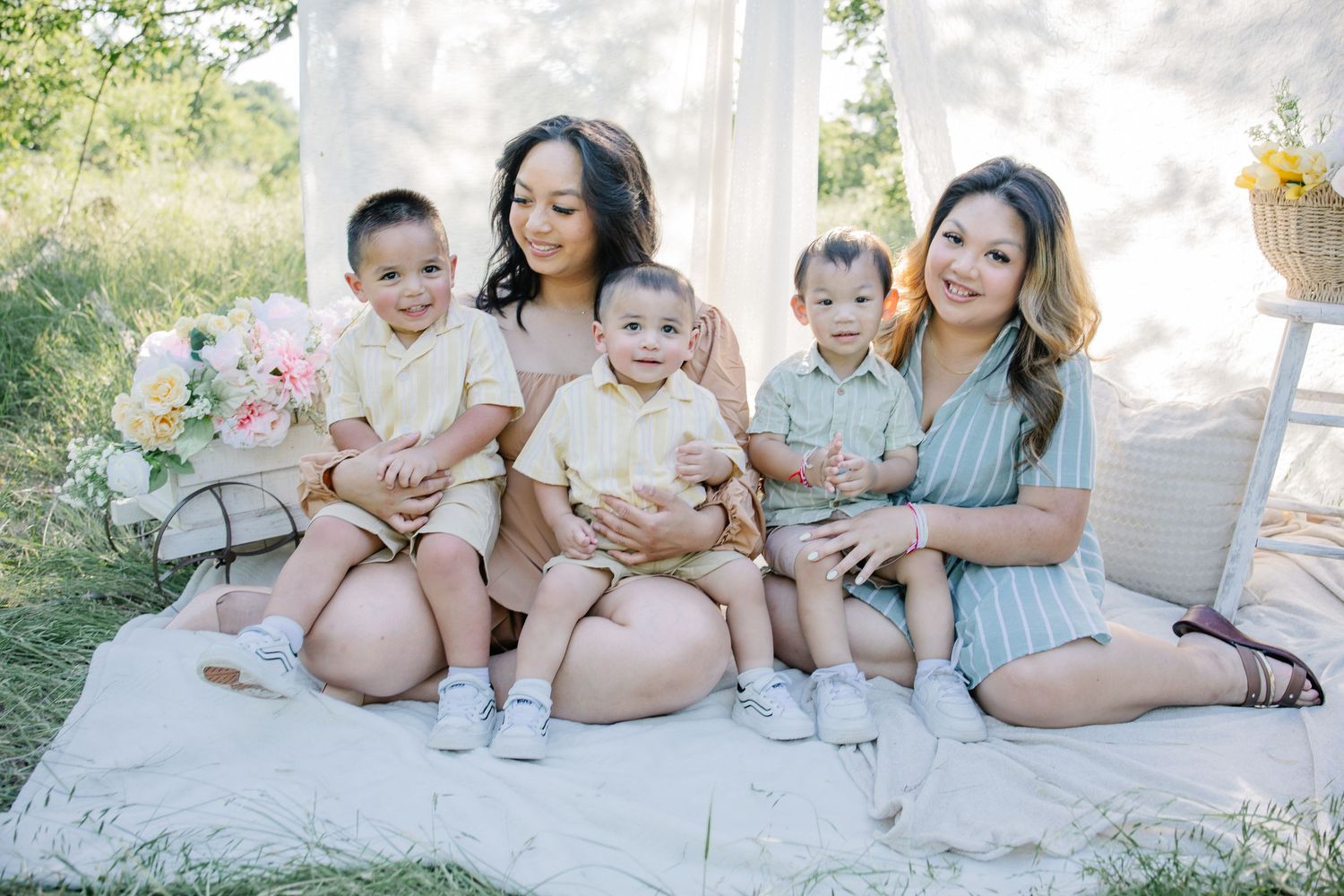 Two women sit outdoors on a blanket with three young toddlers in matching outfits, surrounded by pastel floral decor.